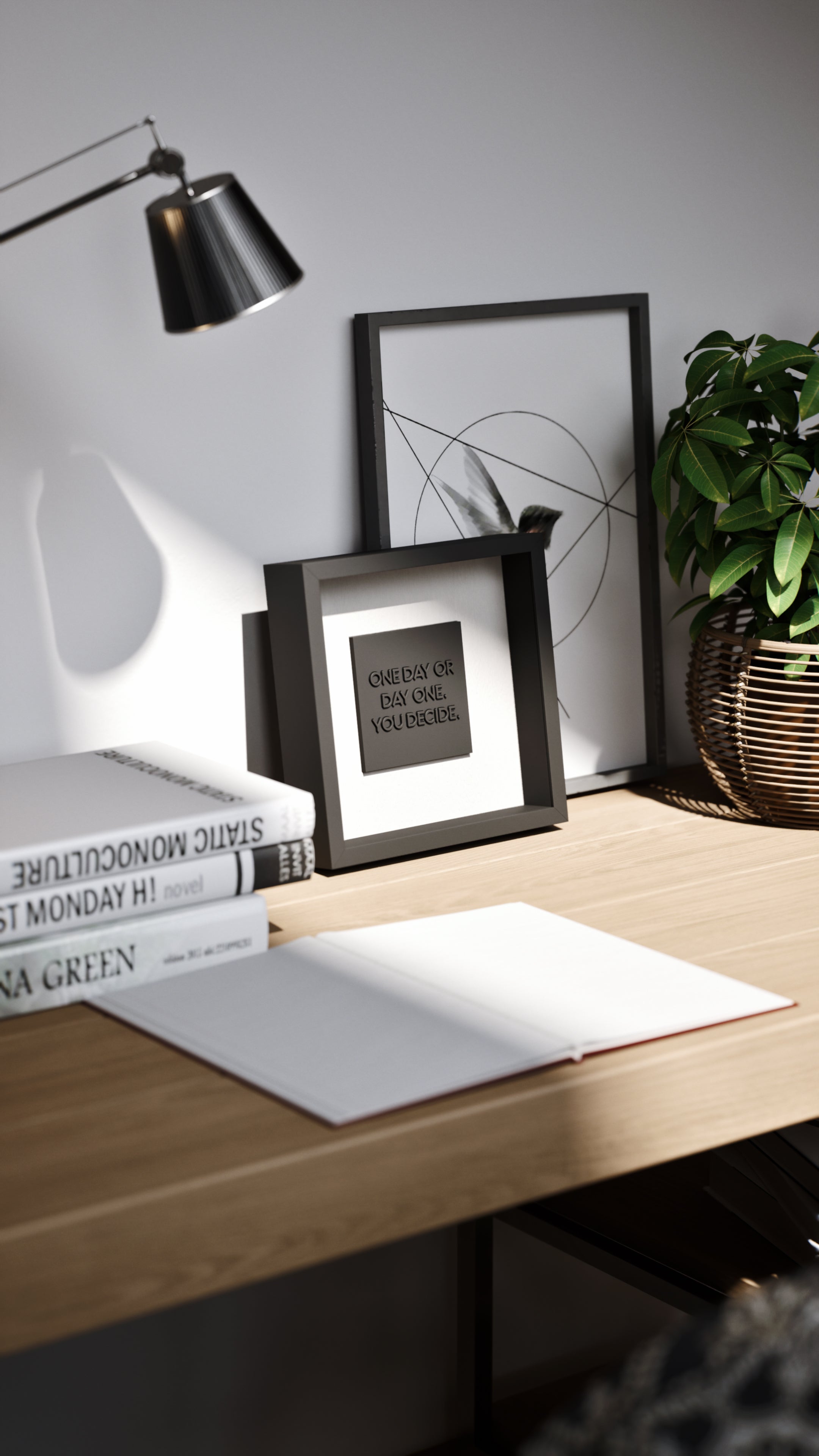 A minimalist desk setup featuring a black shadowbox frame with the motivational quote 'One day or day one. You decide.' The artwork is complemented by neatly stacked books, a wicker plant pot, and geometric wall decor, creating a modern and inspiring workspace.