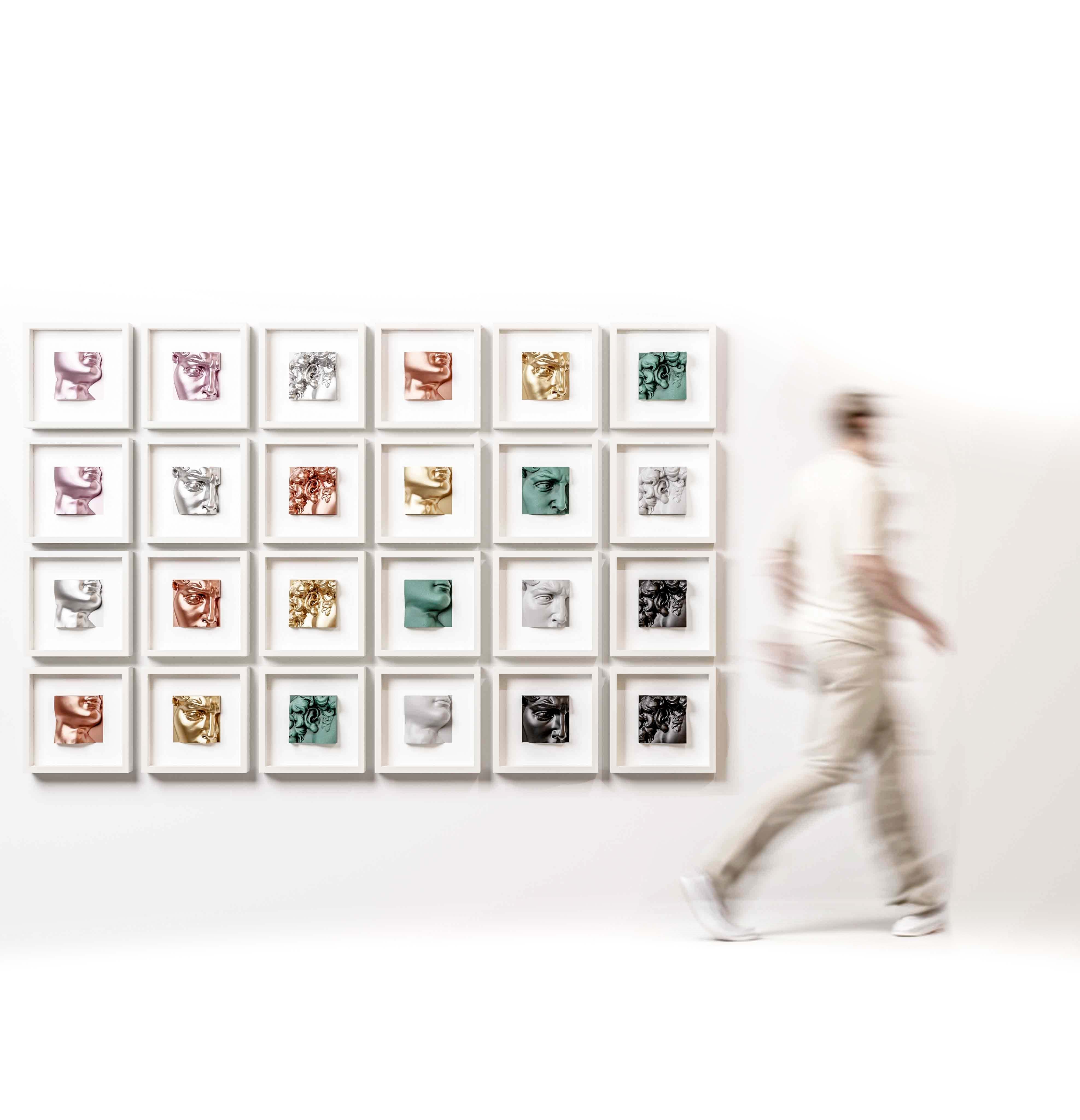 A gallery wall featuring an arrangement of 25 white shadowbox frames, each showcasing a unique sculptural artwork in finishes such as metallic rose, chrome gold, marble white, matte black, and old patina green. The collection creates a visually striking display of textures, forms, and colours, blending classical and modern design elements. A blurred figure walking past adds a dynamic, life-like element to the scene.