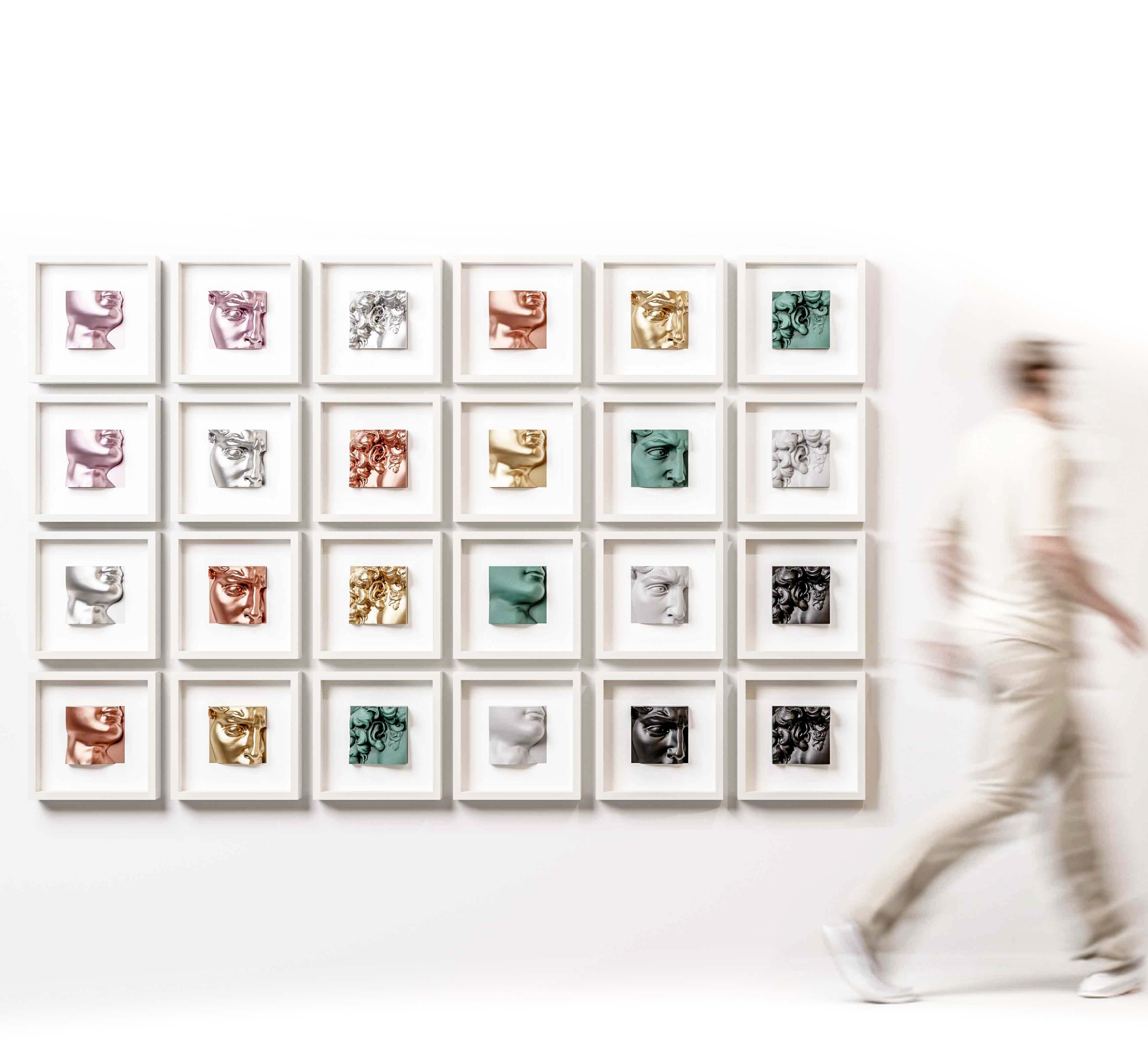 A gallery wall featuring an arrangement of 25 white shadowbox frames, each showcasing a unique sculptural artwork in finishes such as metallic rose, chrome gold, marble white, matte black, and old patina green. The collection creates a visually striking display of textures, forms, and colours, blending classical and modern design elements. A blurred figure walking past adds a dynamic, life-like element to the scene.