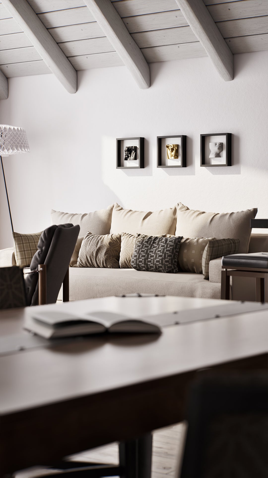 A cozy living room setup with a light beige sofa adorned with patterned cushions, set against a white wall featuring three black shadowbox frames displaying elegant sculptures in black, gold, and white. The space is complemented by a wooden table in the foreground and exposed beam ceilings, creating a warm and inviting atmosphere.