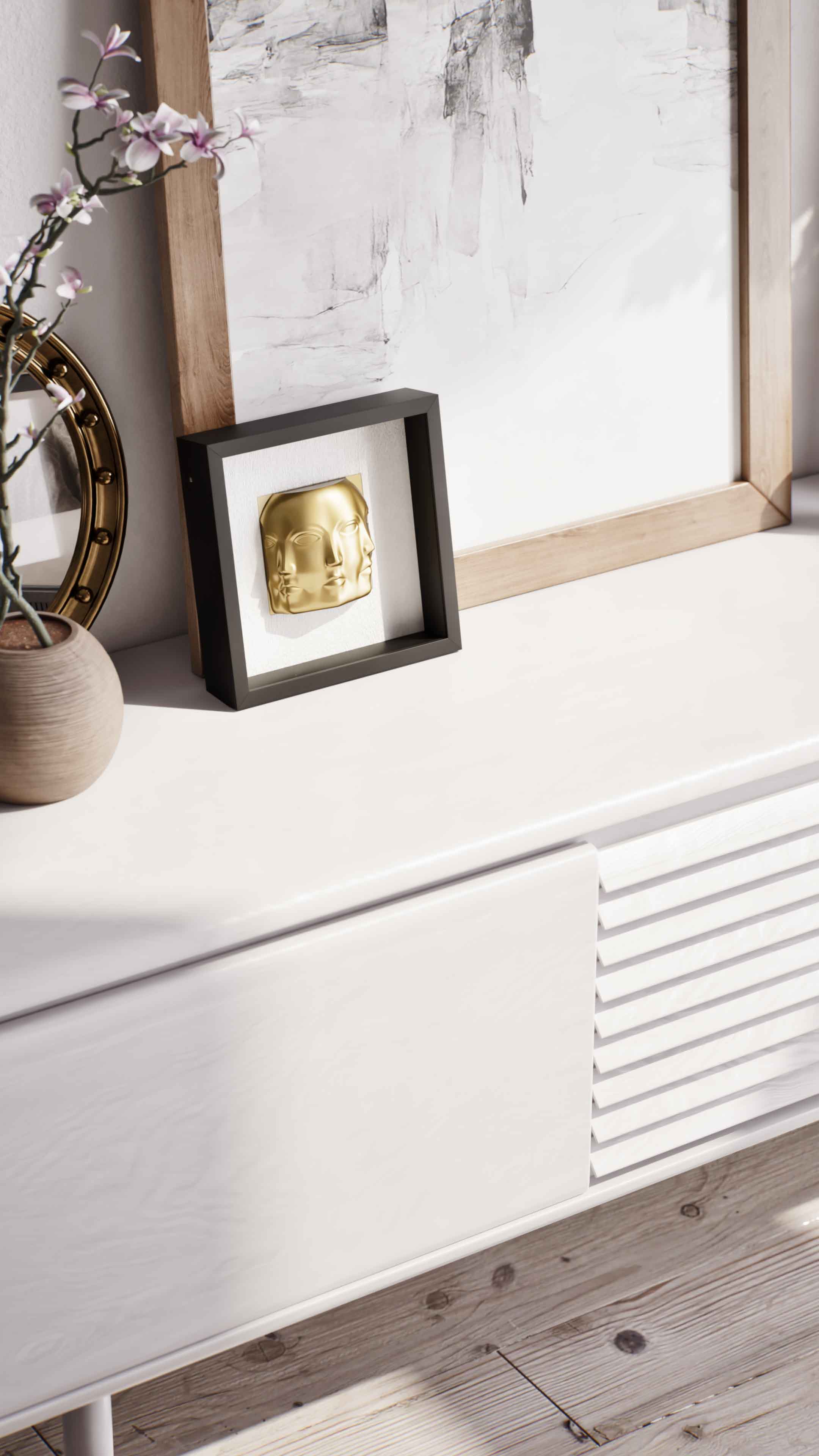 A close-up of a black shadowbox frame featuring a gold three-faced sculpture, displayed on a sleek white cabinet. The setting is complemented by a round, gold-rimmed mirror and a textured wooden picture frame in the background, adding layers of elegance and contemporary style to the composition.