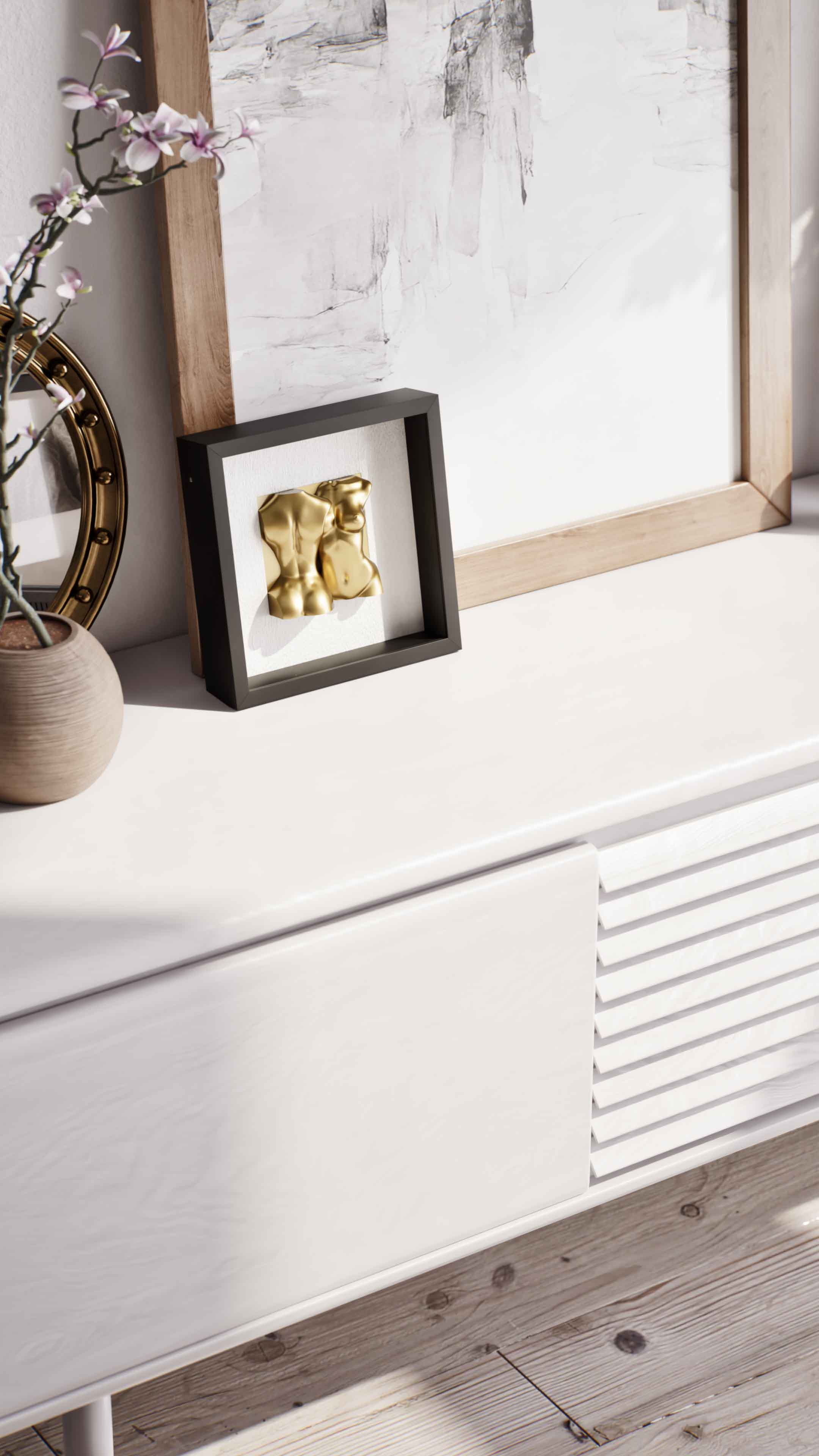 A detailed close-up of a modern white sideboard, showcasing a black shadowbox frame containing a gold sculpture of human torsos. The frame is placed next to a round decorative mirror with a gold rim and a minimalist vase with pink flowering branches, creating an elegant and artistic vignette.