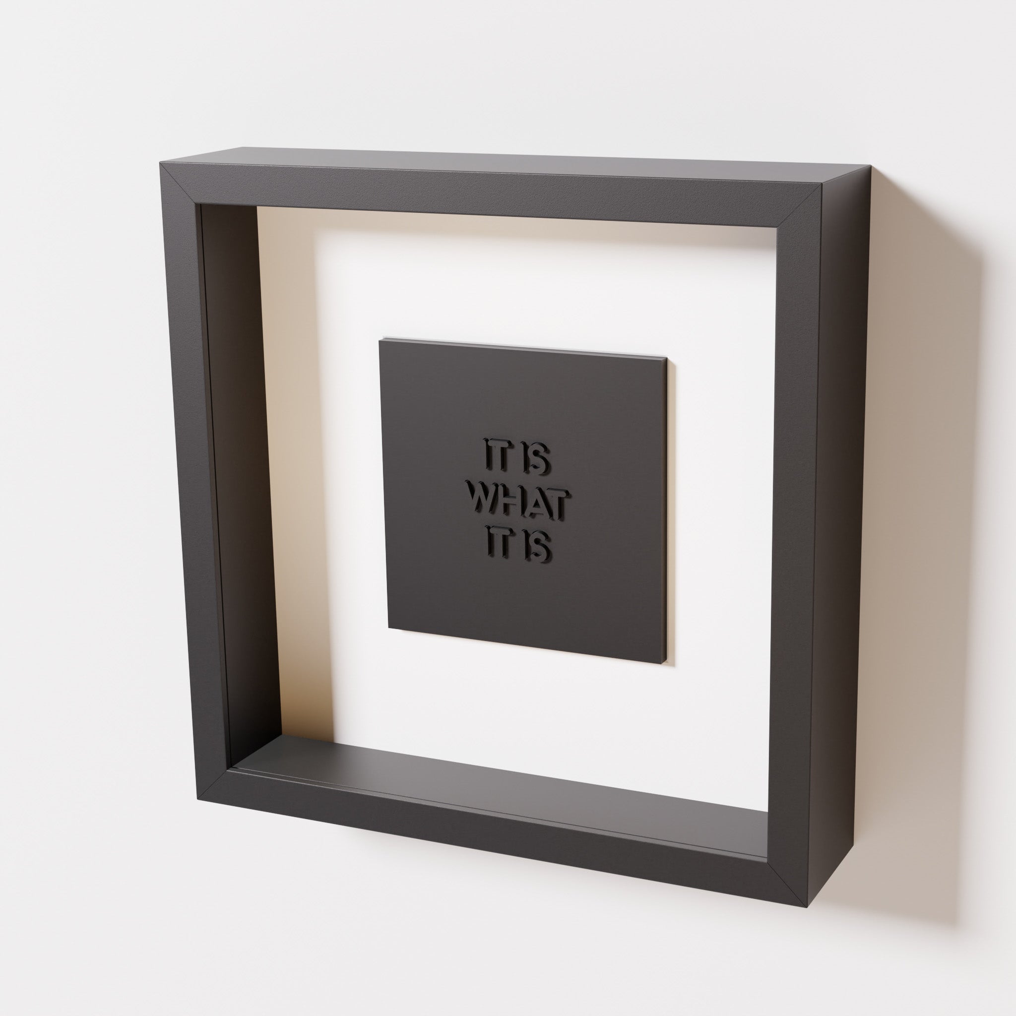 A close-up of a black shadowbox frame containing a matte black artwork featuring the bold phrase 'It Is What It Is' from 'The Chill Pill,' emphasizing its intricate lettering and modern design.