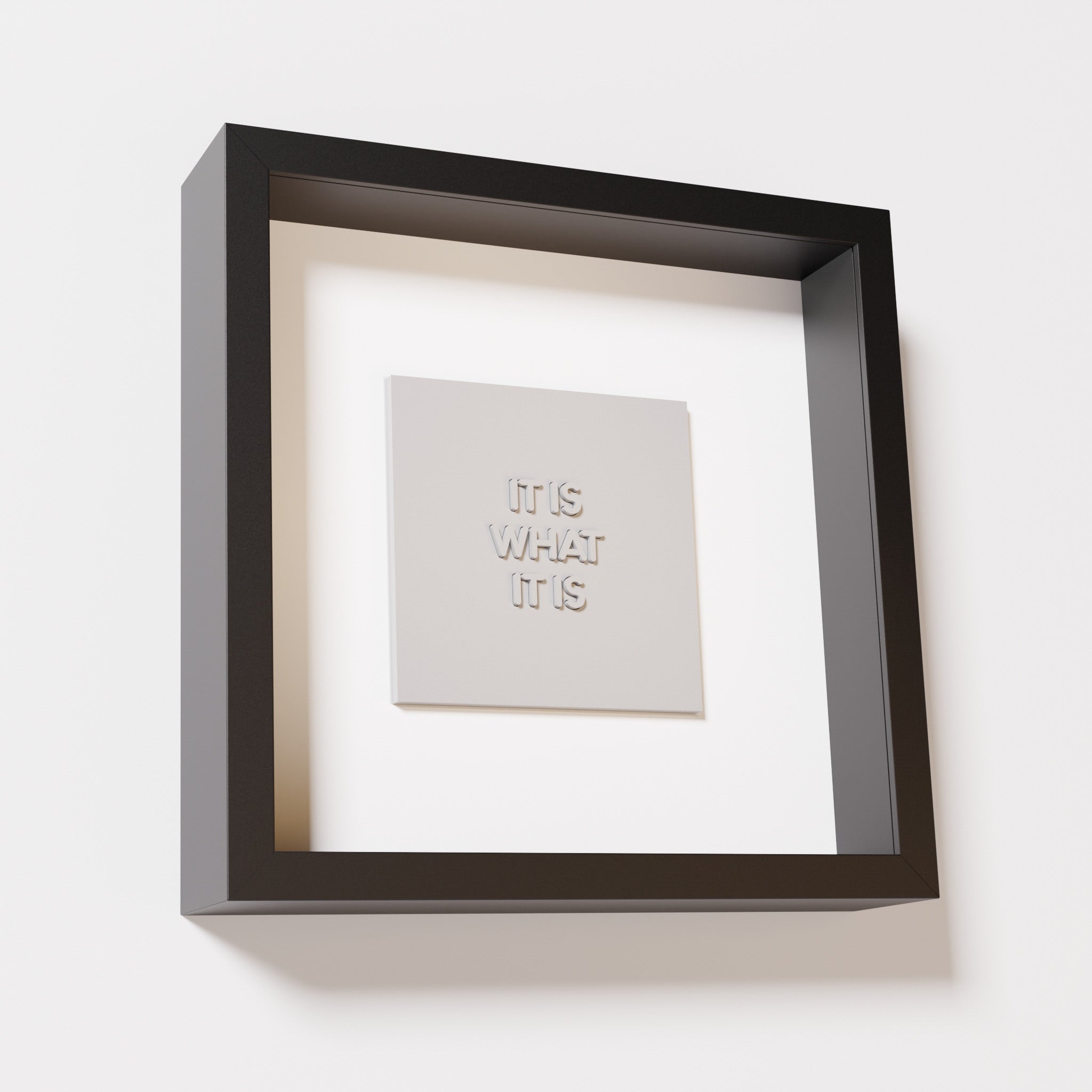 A close-up of a black shadowbox frame containing a marble white artwork featuring the bold phrase 'It Is What It Is' from 'The Chill Pill,' emphasizing its smooth lettering and refined design.