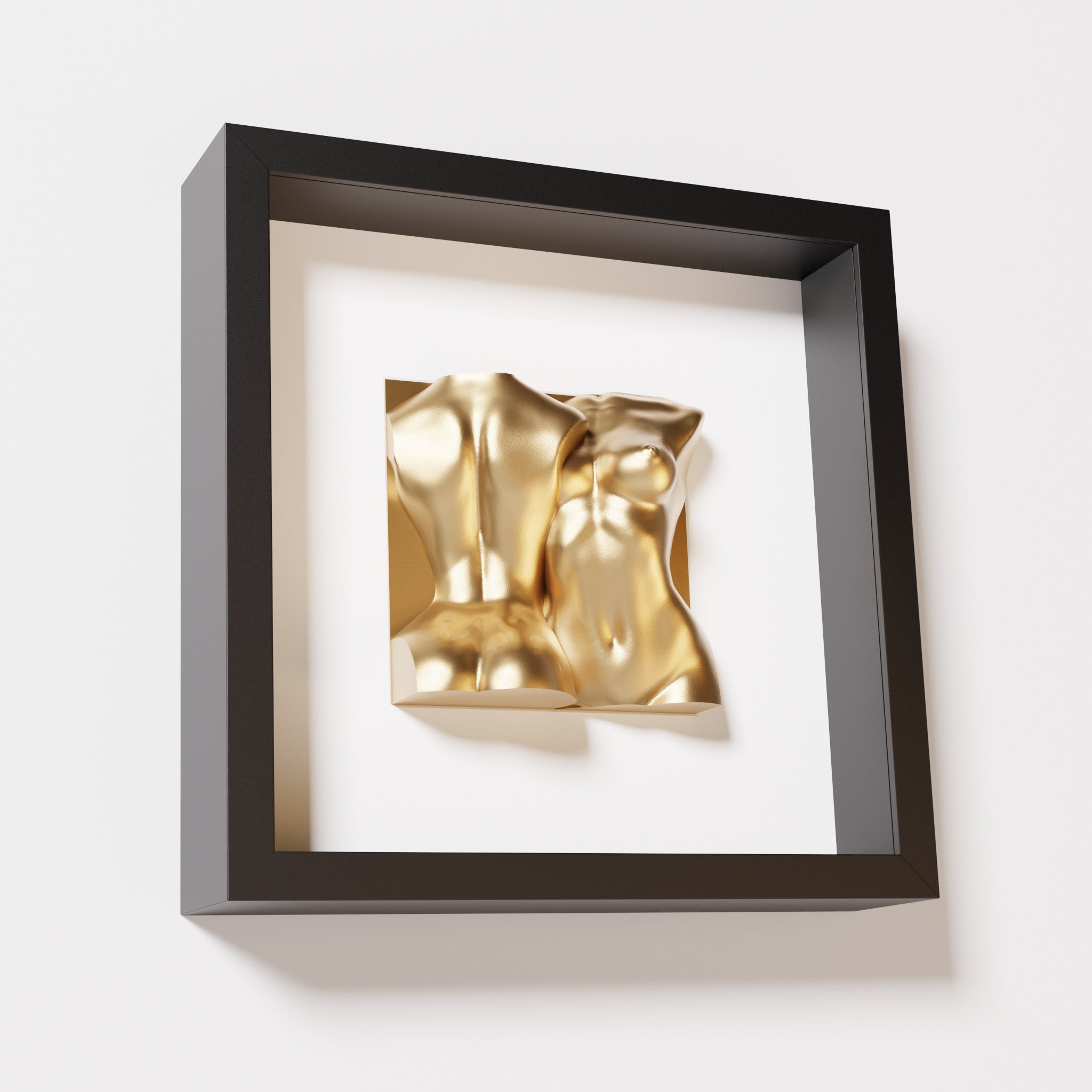 A close-up of a black shadowbox frame holding a chrome gold sculpture of two intertwined female bodies. The reflective gold finish stands out boldly against the sleek black frame for a luxurious effect.