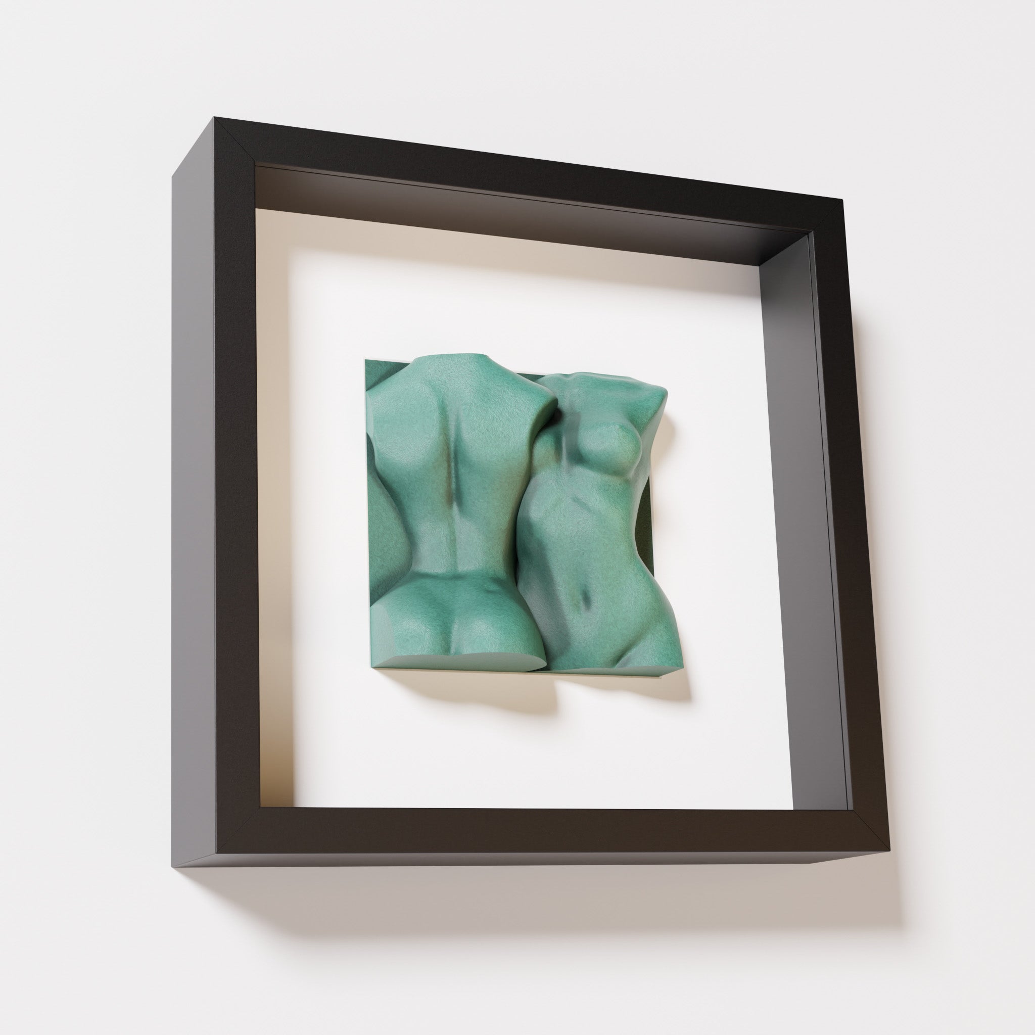 A close-up of a black shadowbox frame containing an old patina sculpture of two intertwined female bodies. The weathered green texture contrasts beautifully with the sleek black frame.