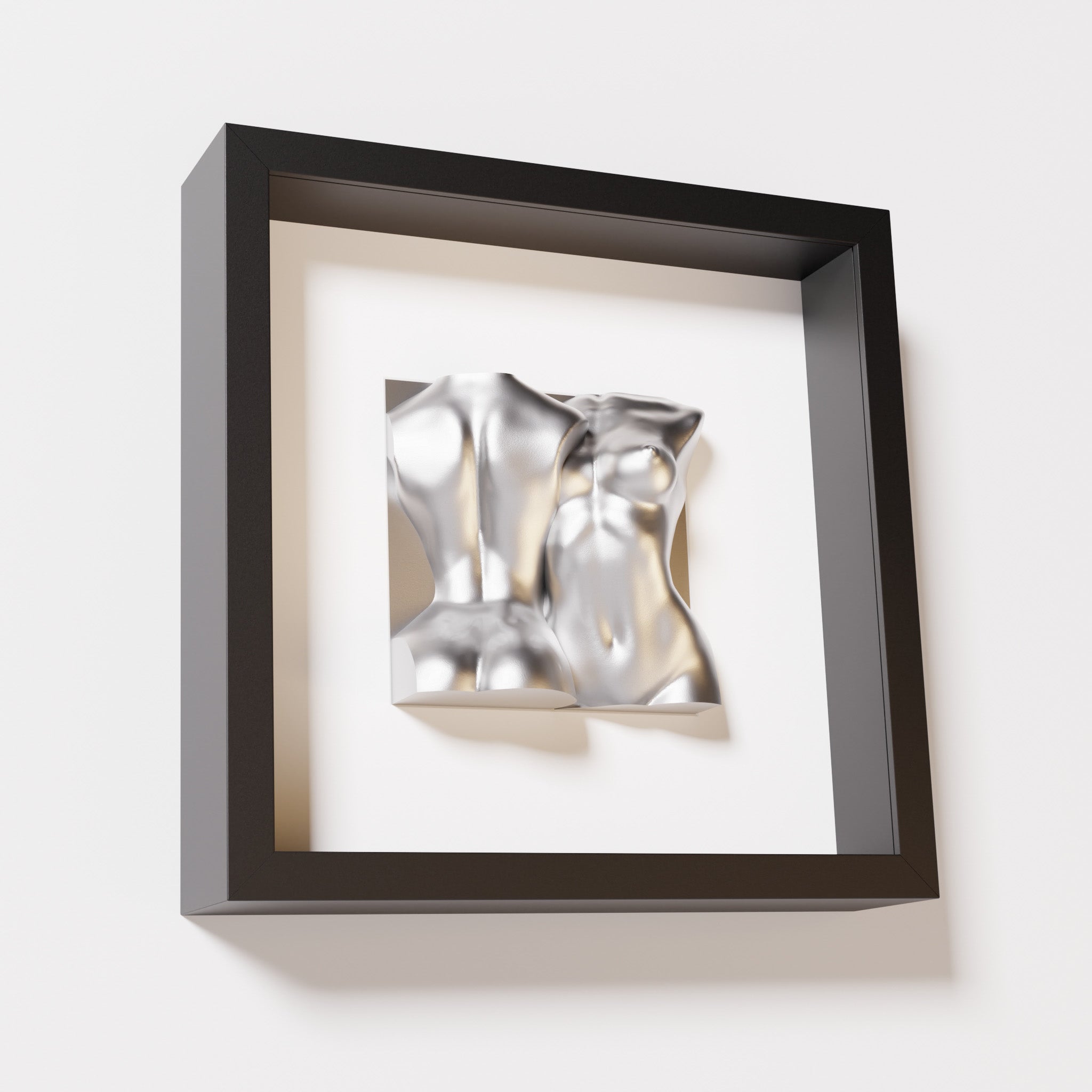 A close-up of a black shadowbox frame holding a chrome silver sculpture of two intertwined female bodies. The reflective silver surface creates a bold contrast against the dark frame.