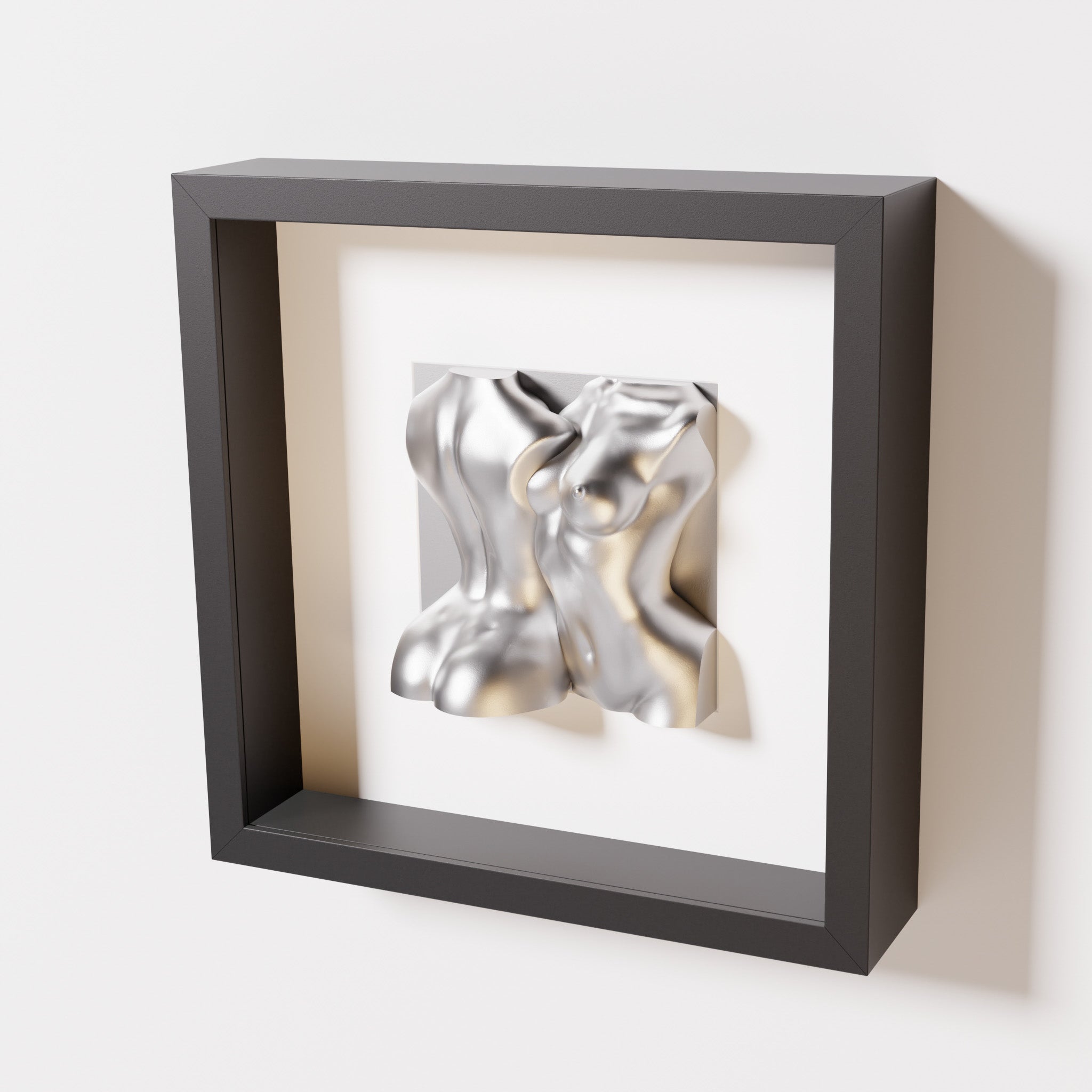 A close-up of a black shadowbox frame holding a chrome silver sculpture of two intertwined female bodies. The reflective silver surface creates a bold contrast against the dark frame.