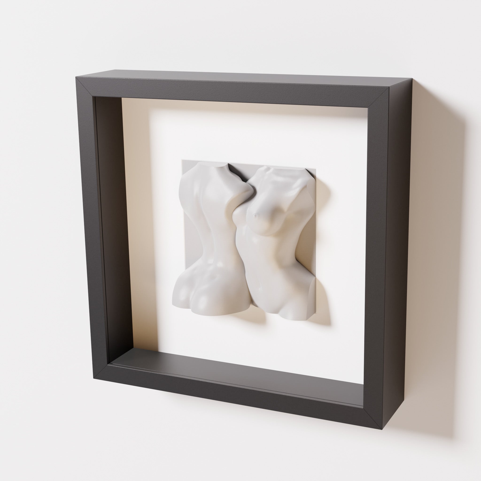 A close-up of a black shadowbox frame containing a marble white sculpture of two intertwined female bodies. The smooth white figures stand out boldly against the dark frame.