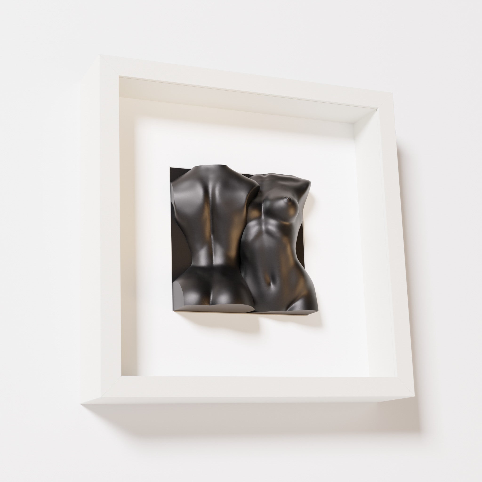 A close-up of a white shadowbox frame containing a matte black sculpture depicting two intertwined female bodies. The smooth, abstract forms of the figures contrast elegantly with the clean white frame.