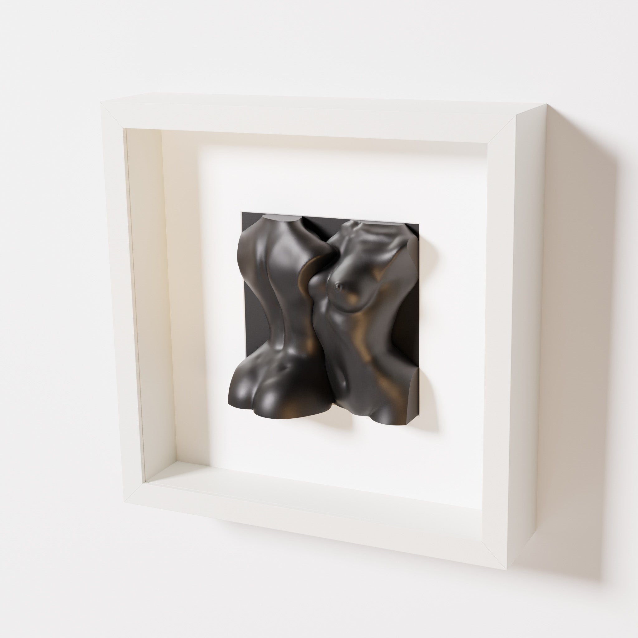 A close-up of a white shadowbox frame containing a matte black sculpture depicting two intertwined female bodies. The smooth, abstract forms of the figures contrast elegantly with the clean white frame.