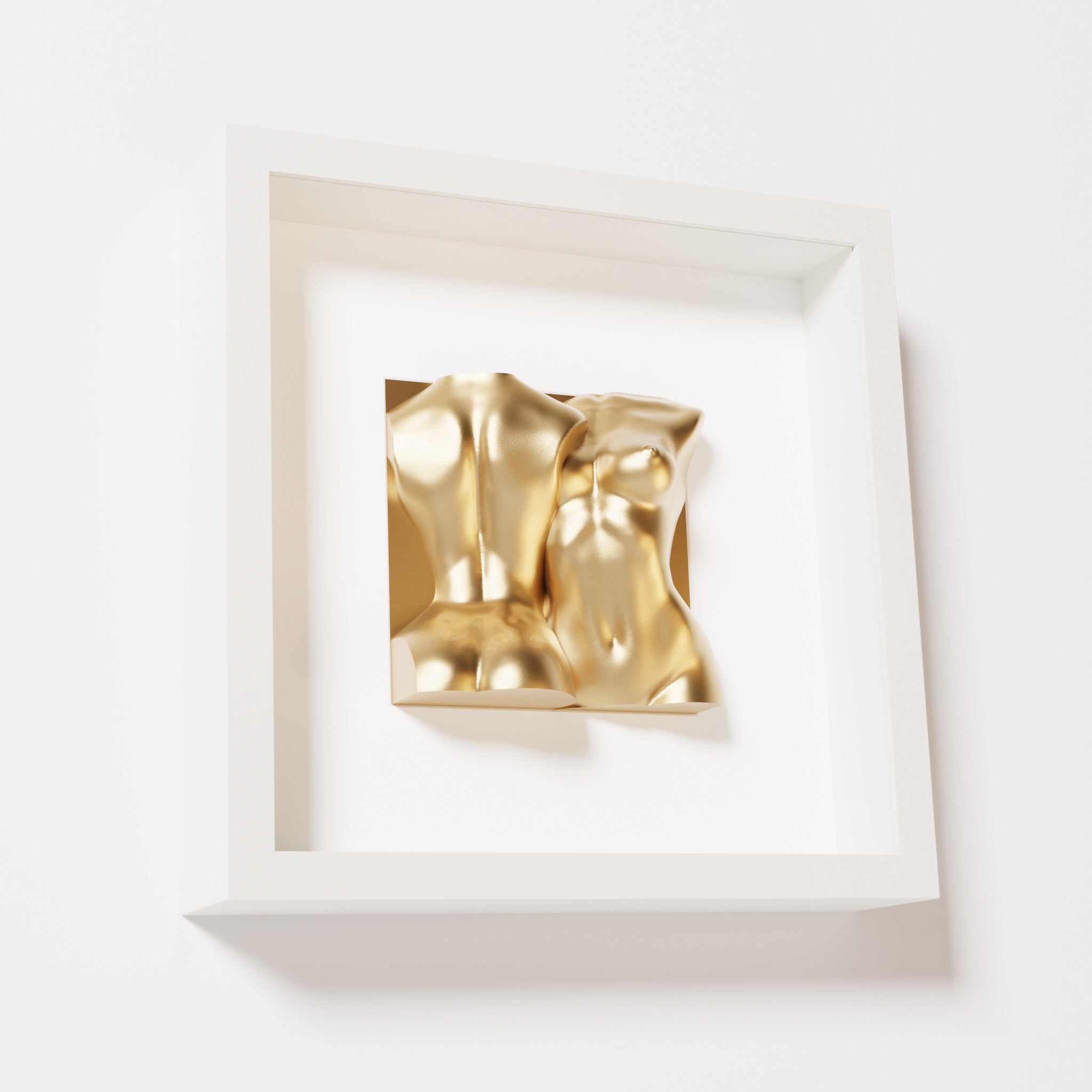 A close-up of a white shadowbox frame containing a chrome gold sculpture of two intertwined female bodies. The reflective gold surface adds a sense of opulence to the smooth, abstract forms.