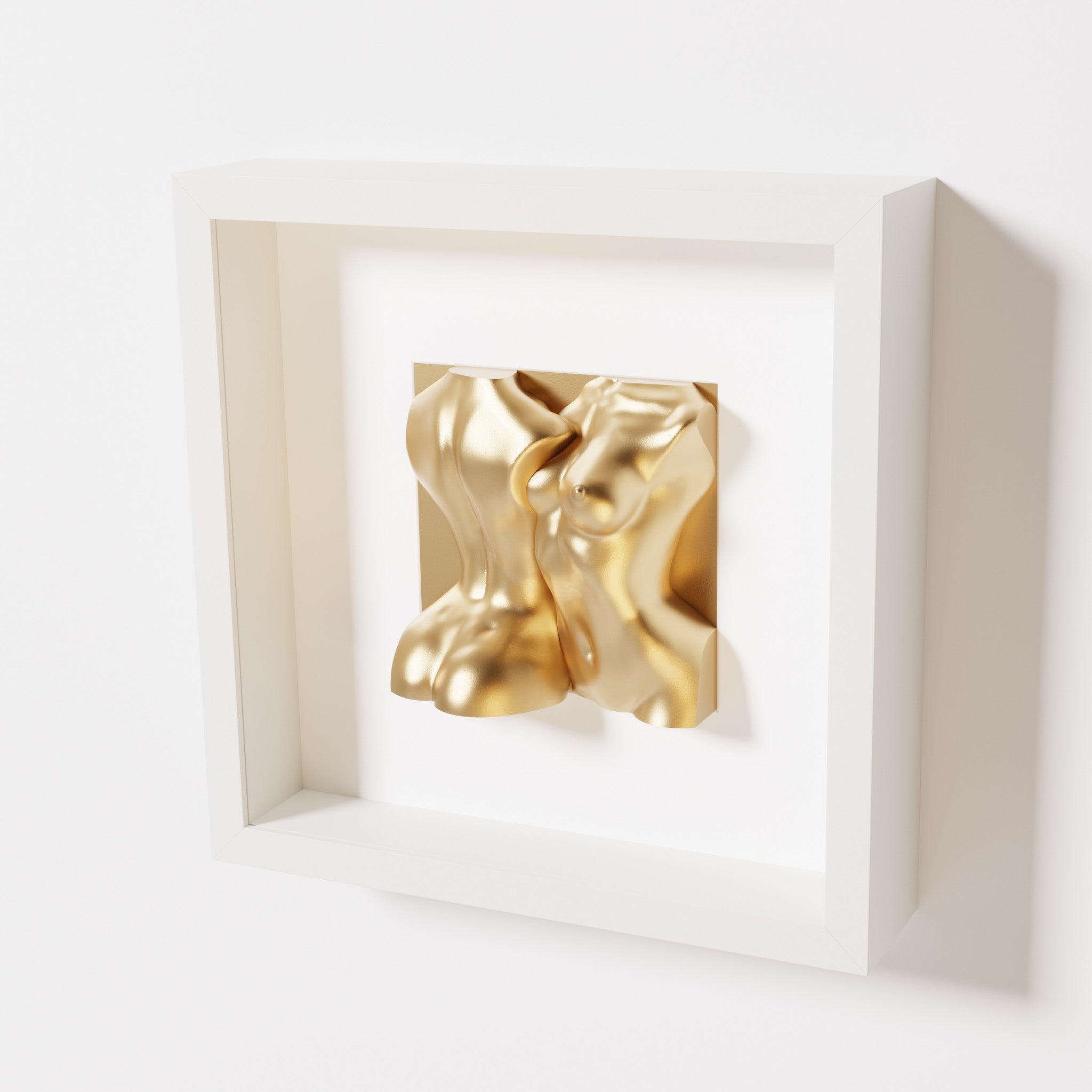 A close-up of a white shadowbox frame containing a chrome gold sculpture of two intertwined female bodies. The reflective gold surface adds a sense of opulence to the smooth, abstract forms.