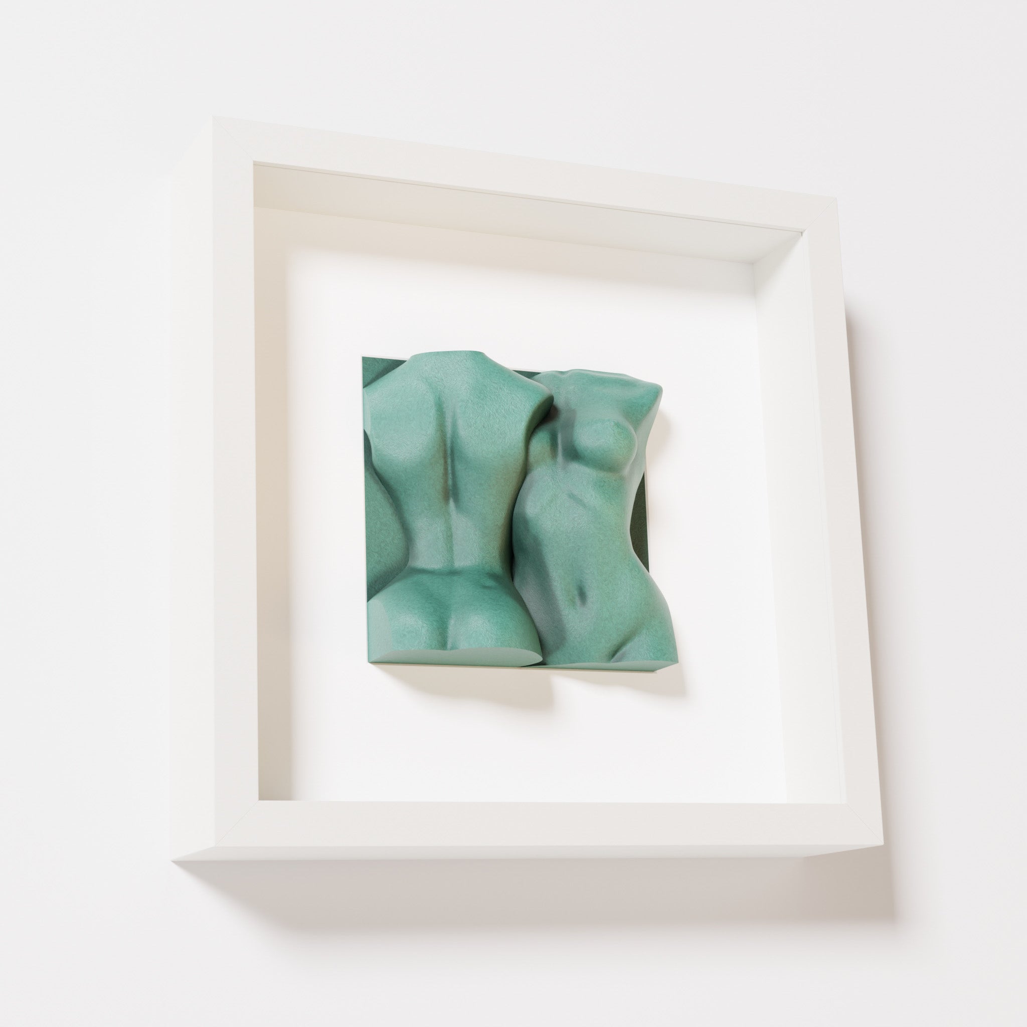 A close-up of a white shadowbox frame containing an old patina sculpture of two intertwined female bodies. The textured, weathered green finish lends a classical and timeless character to the artwork.