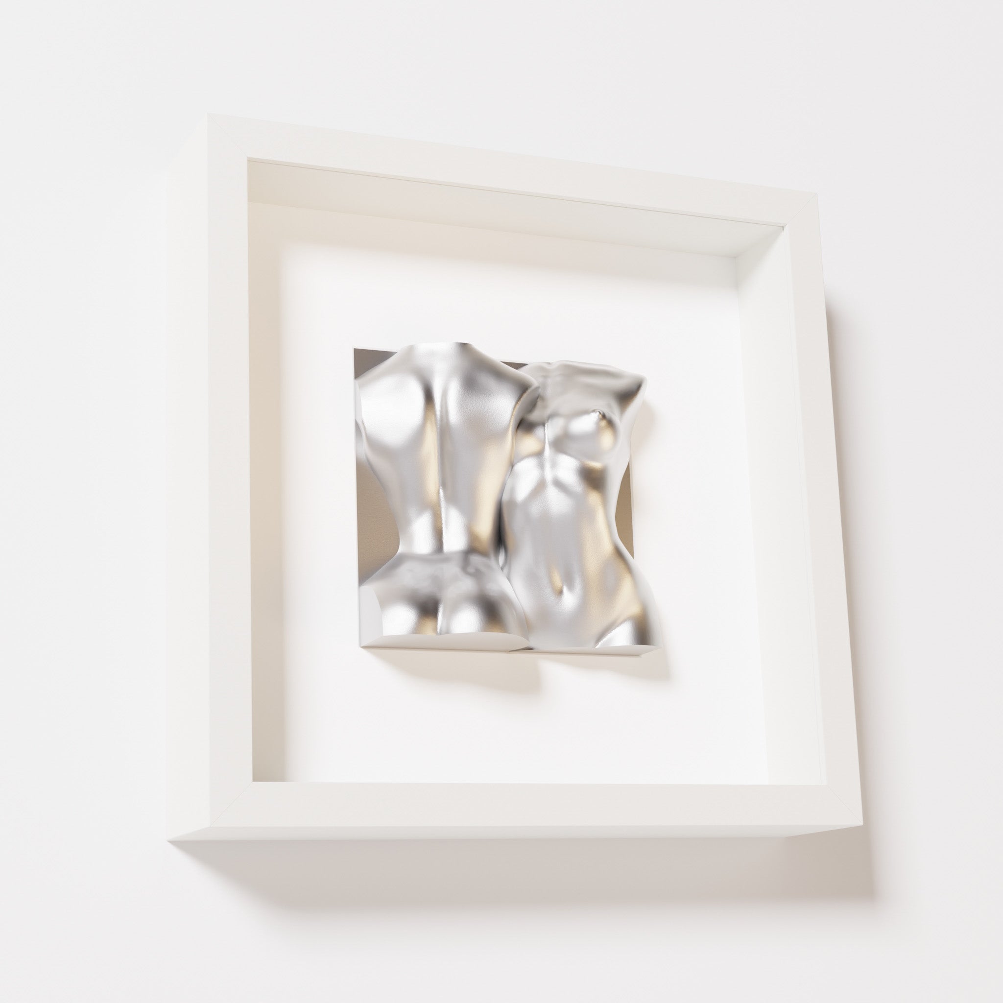 A close-up of a white shadowbox frame containing a chrome silver sculpture of two intertwined female bodies. The reflective metallic surface highlights the fluid, modern forms of the artwork.