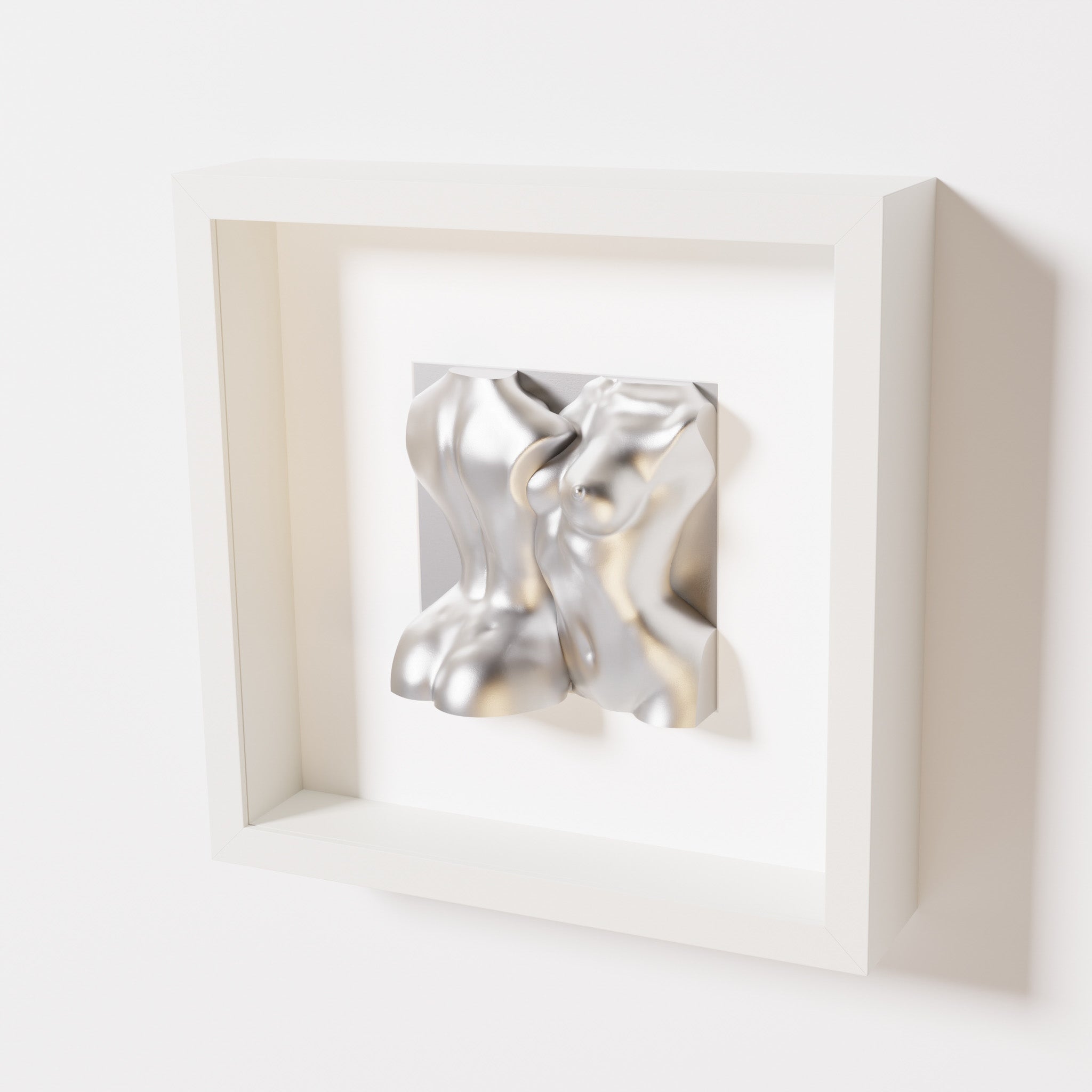 A close-up of a white shadowbox frame containing a chrome silver sculpture of two intertwined female bodies. The reflective metallic surface highlights the fluid, modern forms of the artwork.