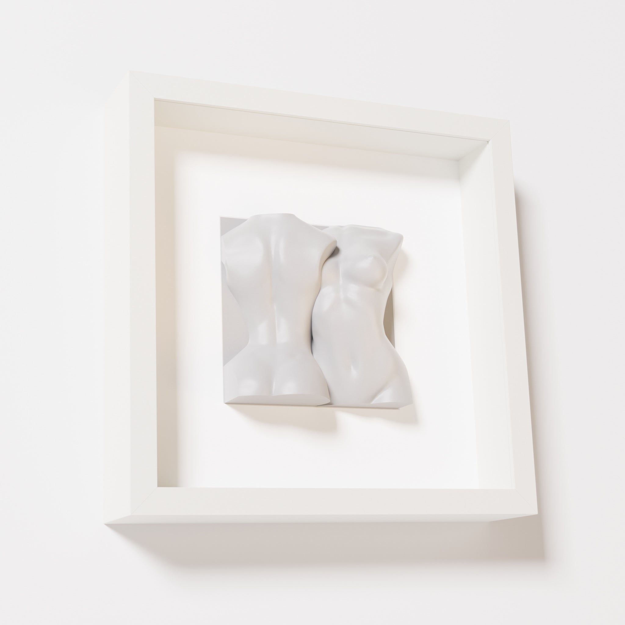 A close-up of a white shadowbox frame containing a marble white sculpture depicting two intertwined female bodies. The soft, fluid forms of the figures create a timeless and elegant aesthetic.