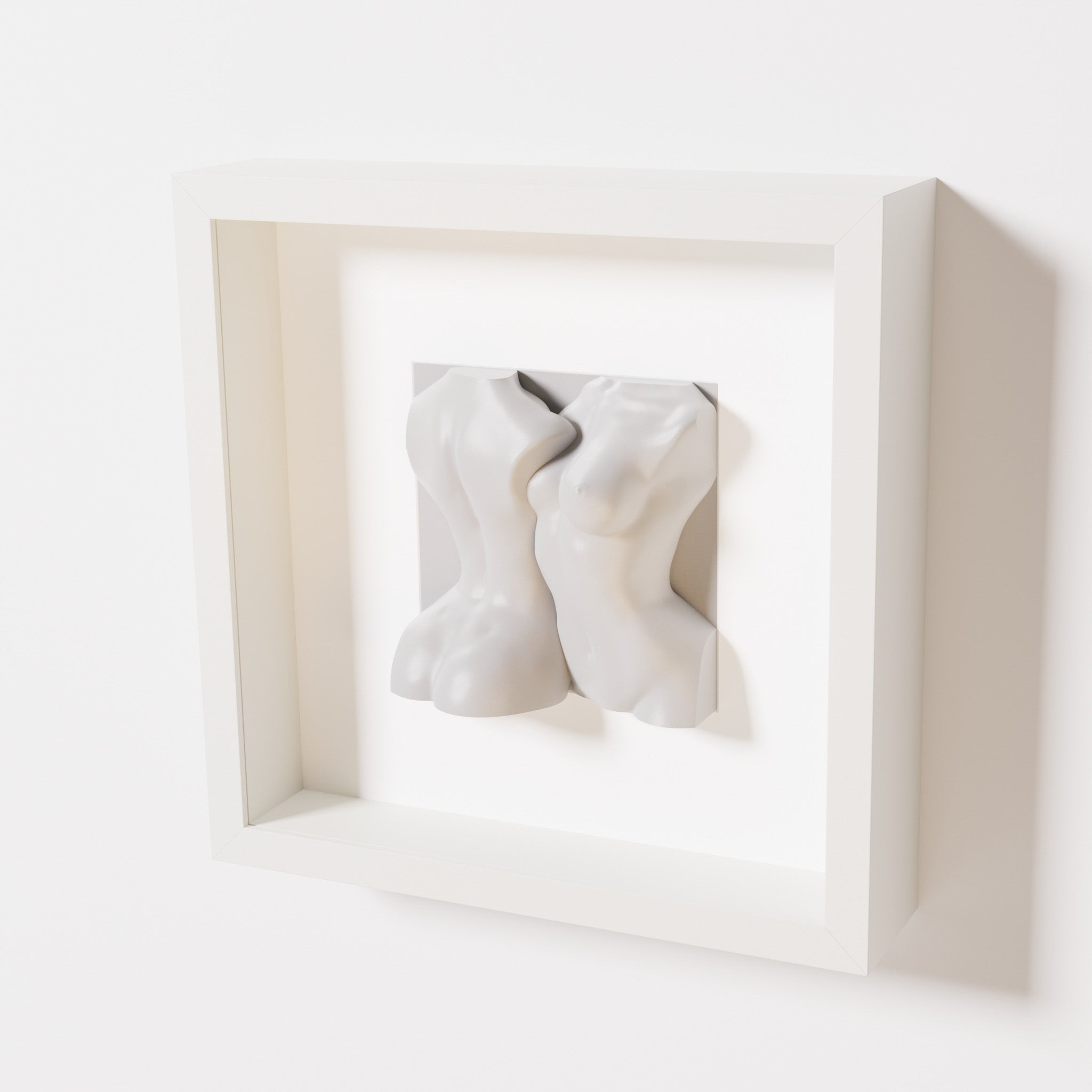 A close-up of a white shadowbox frame containing a marble white sculpture depicting two intertwined female bodies. The soft, fluid forms of the figures create a timeless and elegant aesthetic.