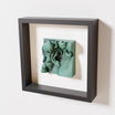 A close-up of a black shadowbox frame featuring an old patina sculpture of 'David's Ear.' The green textured finish and intricate curls stand out against the dark frame.