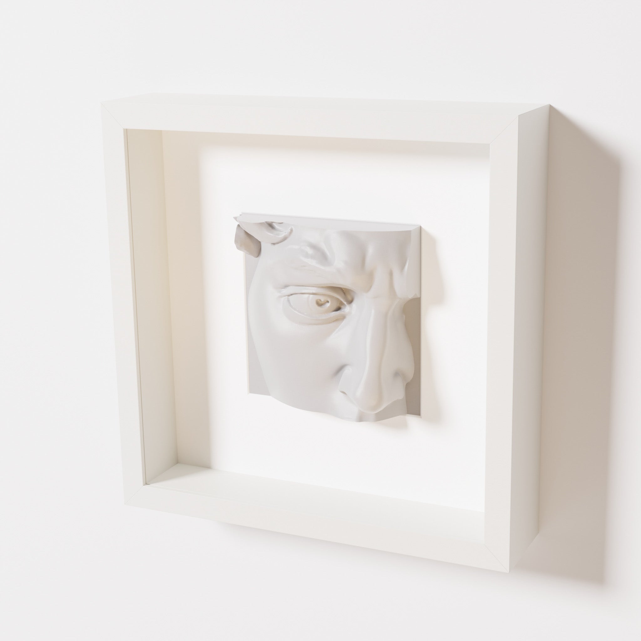 A close-up of a white shadowbox frame containing a marble white sculpture of 'David's Eye,' highlighting the intricate details of the eye, brow, and surrounding textures.