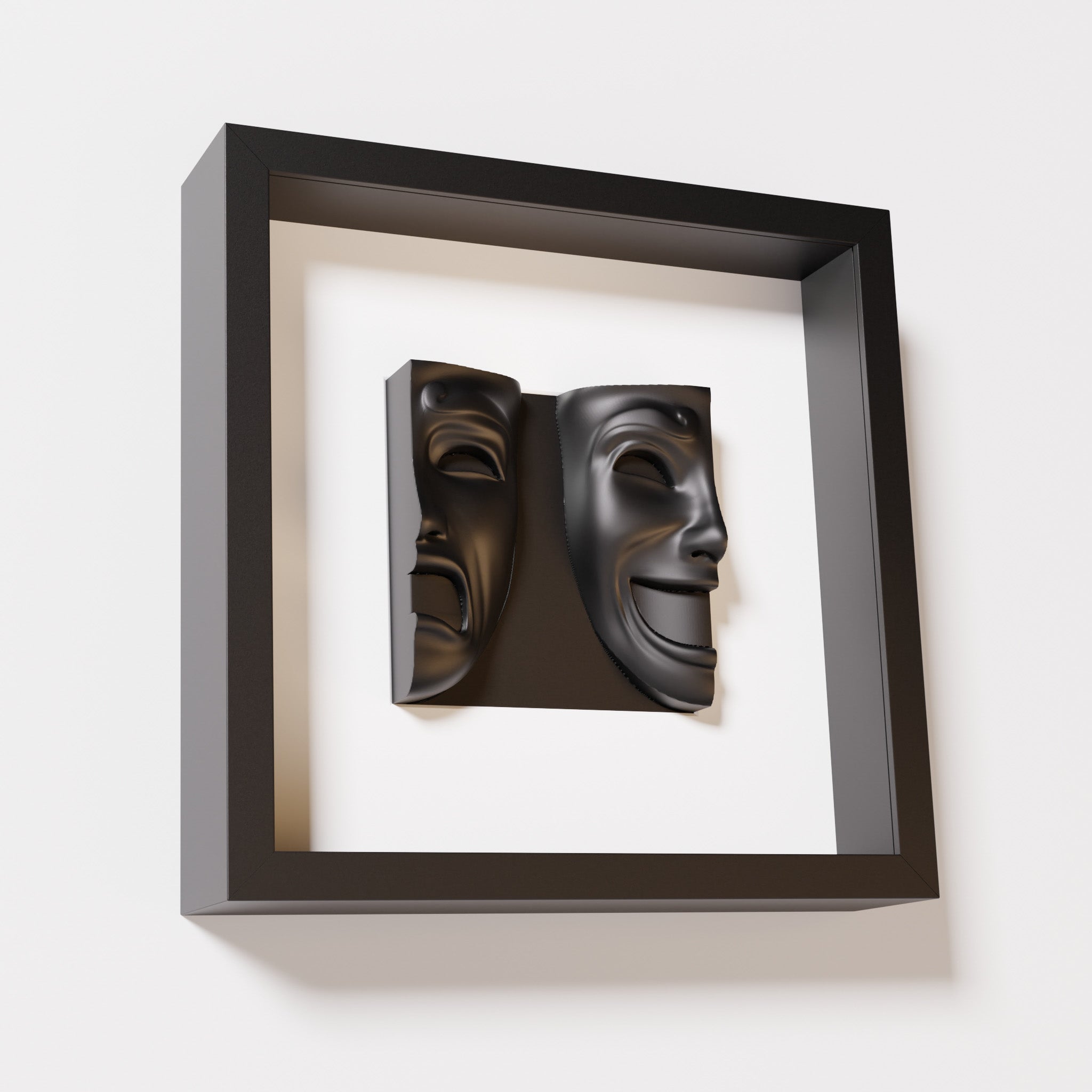 A close-up of a black shadowbox frame containing a matte black sculpture of 'Despair & Joy,' showcasing two expressive masks side by side: one with a sorrowful expression and the other with a joyful smile.