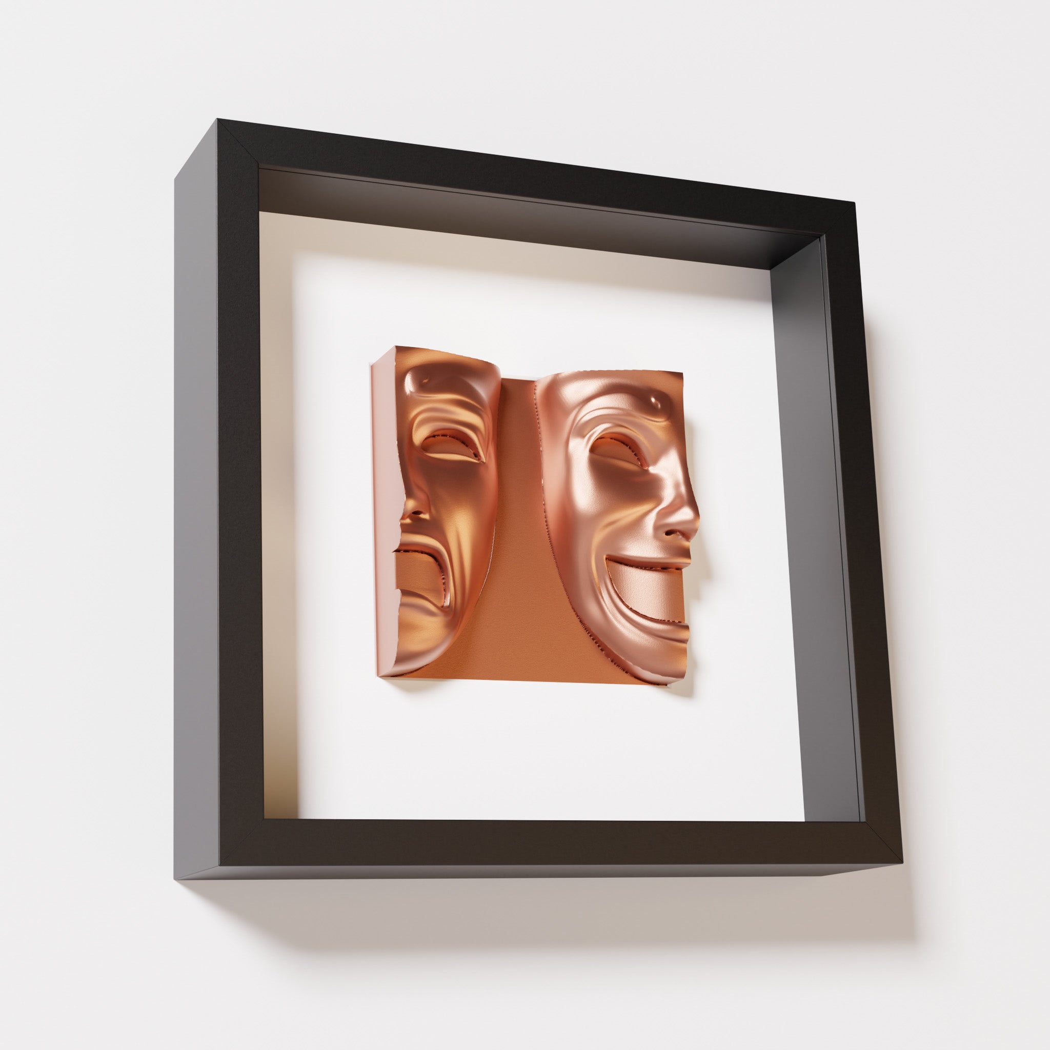 A close-up of a black shadowbox frame containing a chrome copper sculpture of 'Despair & Joy,' showcasing two expressive masks side by side: one with a sorrowful expression and the other with a joyful smile.