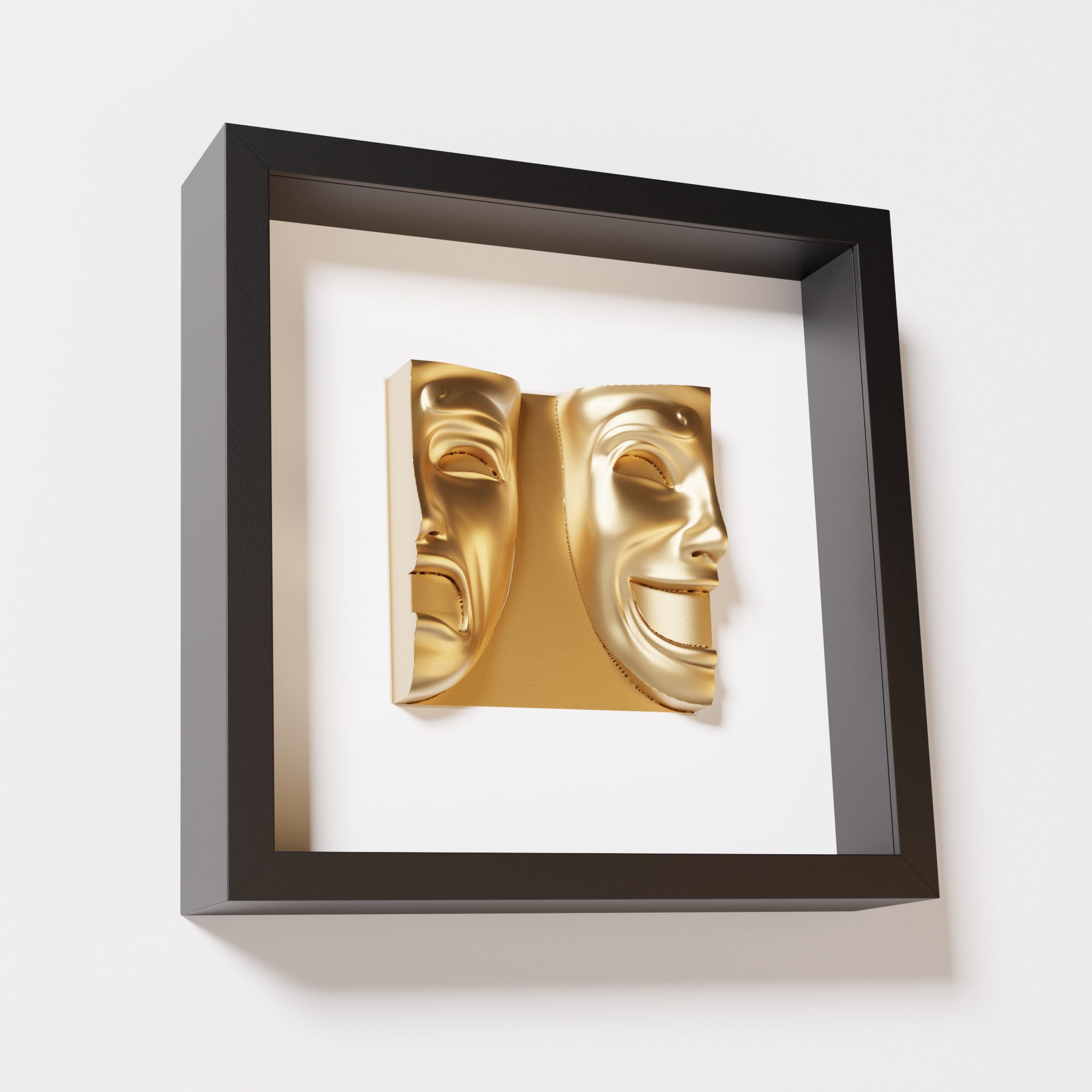 A close-up of a black shadowbox frame containing a chrome gold sculpture of 'Despair & Joy,' showcasing two expressive masks side by side: one with a sorrowful expression and the other with a joyful smile.