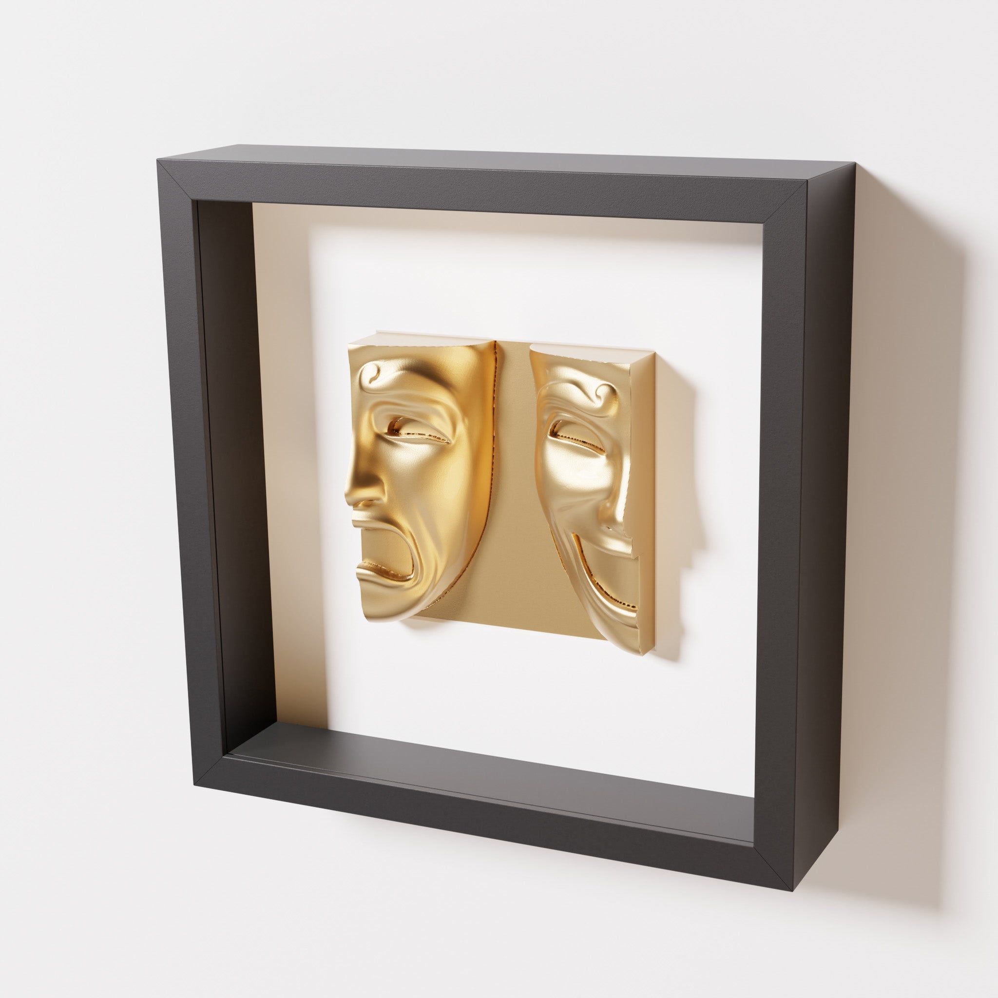 A close-up of a black shadowbox frame containing a chrome gold sculpture of 'Despair & Joy,' showcasing two expressive masks side by side: one with a sorrowful expression and the other with a joyful smile.