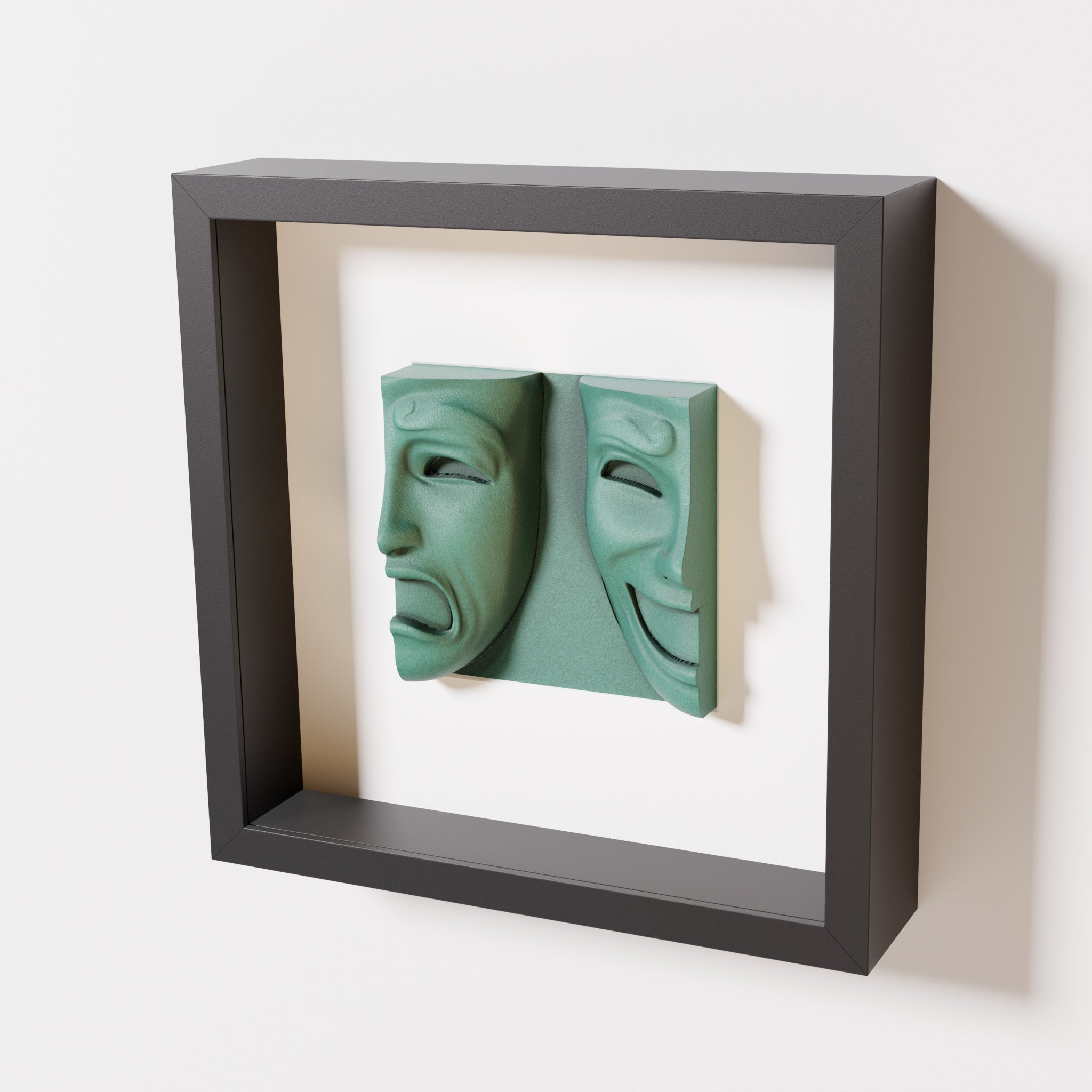 A close-up of a black shadowbox frame containing an old patina sculpture of 'Despair & Joy,' showcasing two expressive masks side by side: one with a sorrowful expression and the other with a joyful smile.