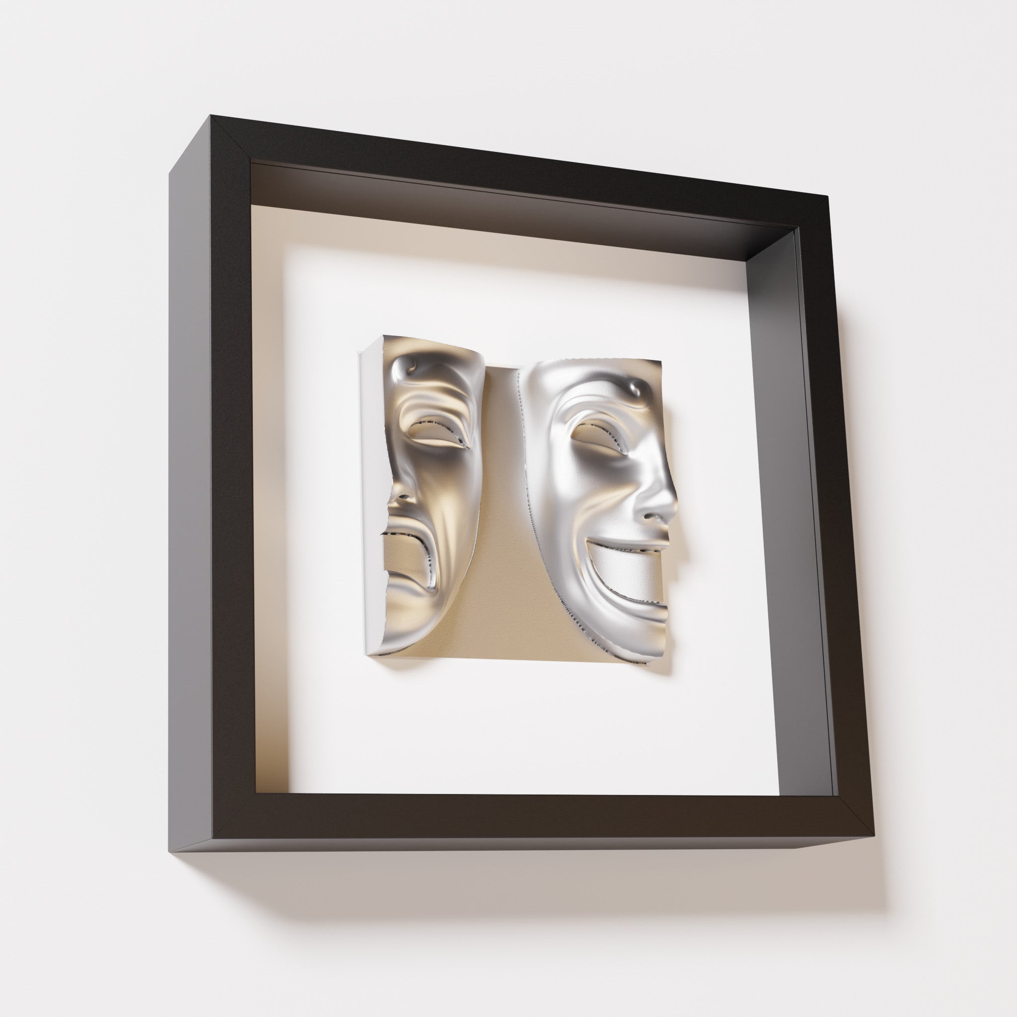 A close-up of a black shadowbox frame containing a chrome silver sculpture of 'Despair & Joy,' showcasing two expressive masks side by side: one with a sorrowful expression and the other with a joyful smile.