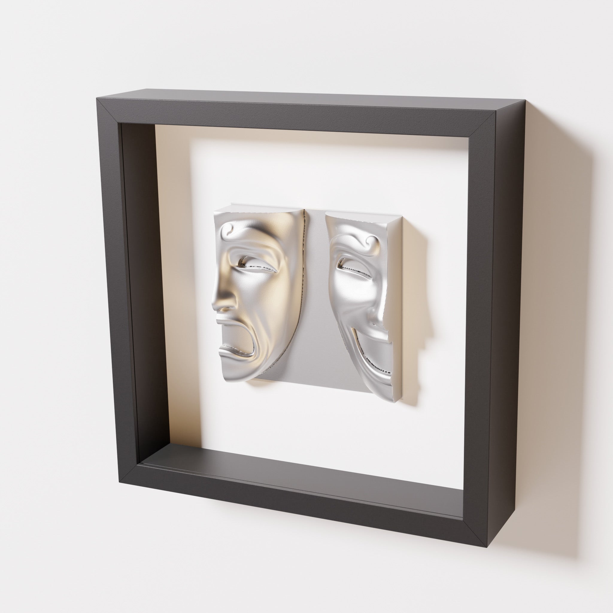 A close-up of a black shadowbox frame containing a chrome silver sculpture of 'Despair & Joy,' showcasing two expressive masks side by side: one with a sorrowful expression and the other with a joyful smile.