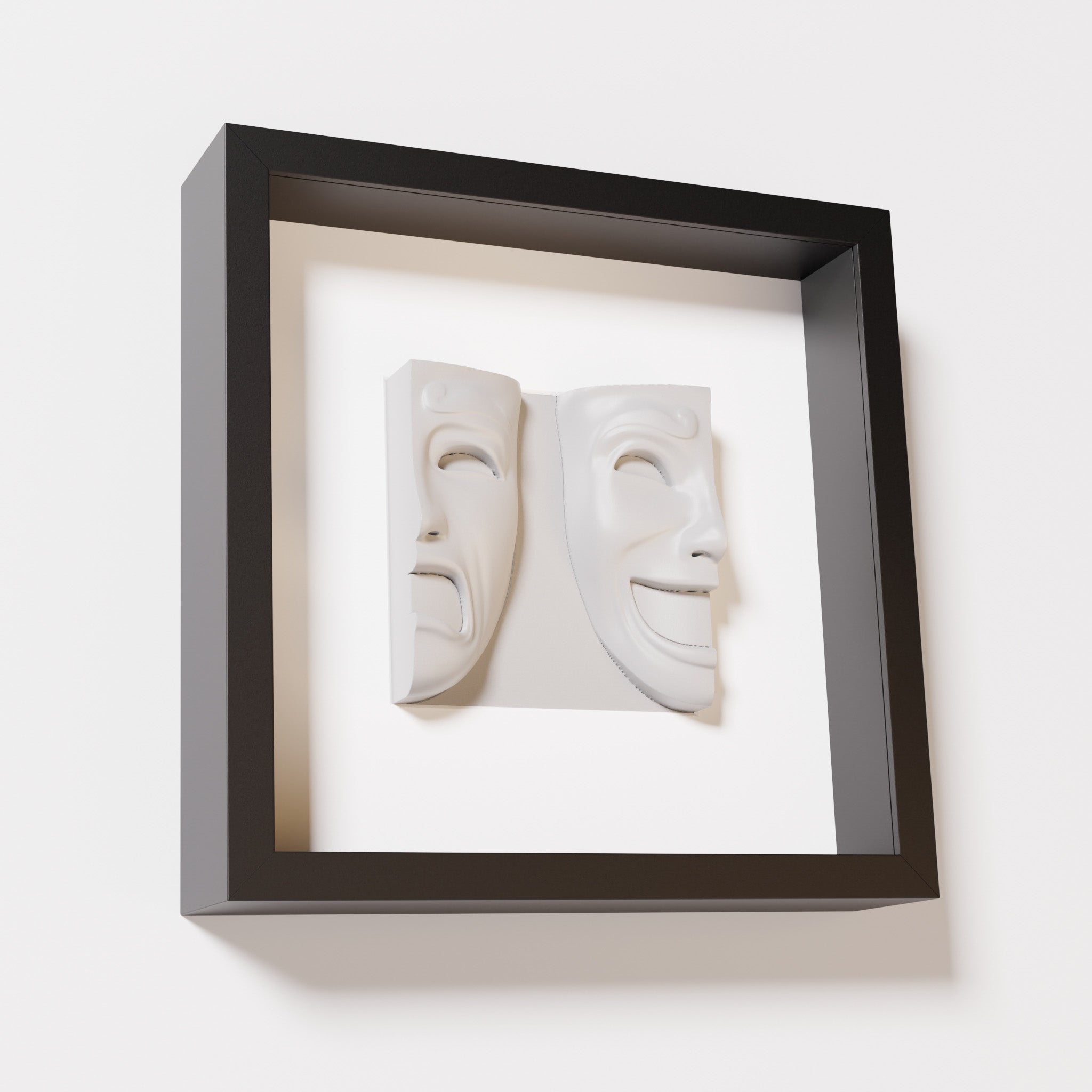 A close-up of a black shadowbox frame containing a marble white sculpture of 'Despair & Joy,' showcasing two expressive masks side by side: one with a sorrowful expression and the other with a joyful smile.