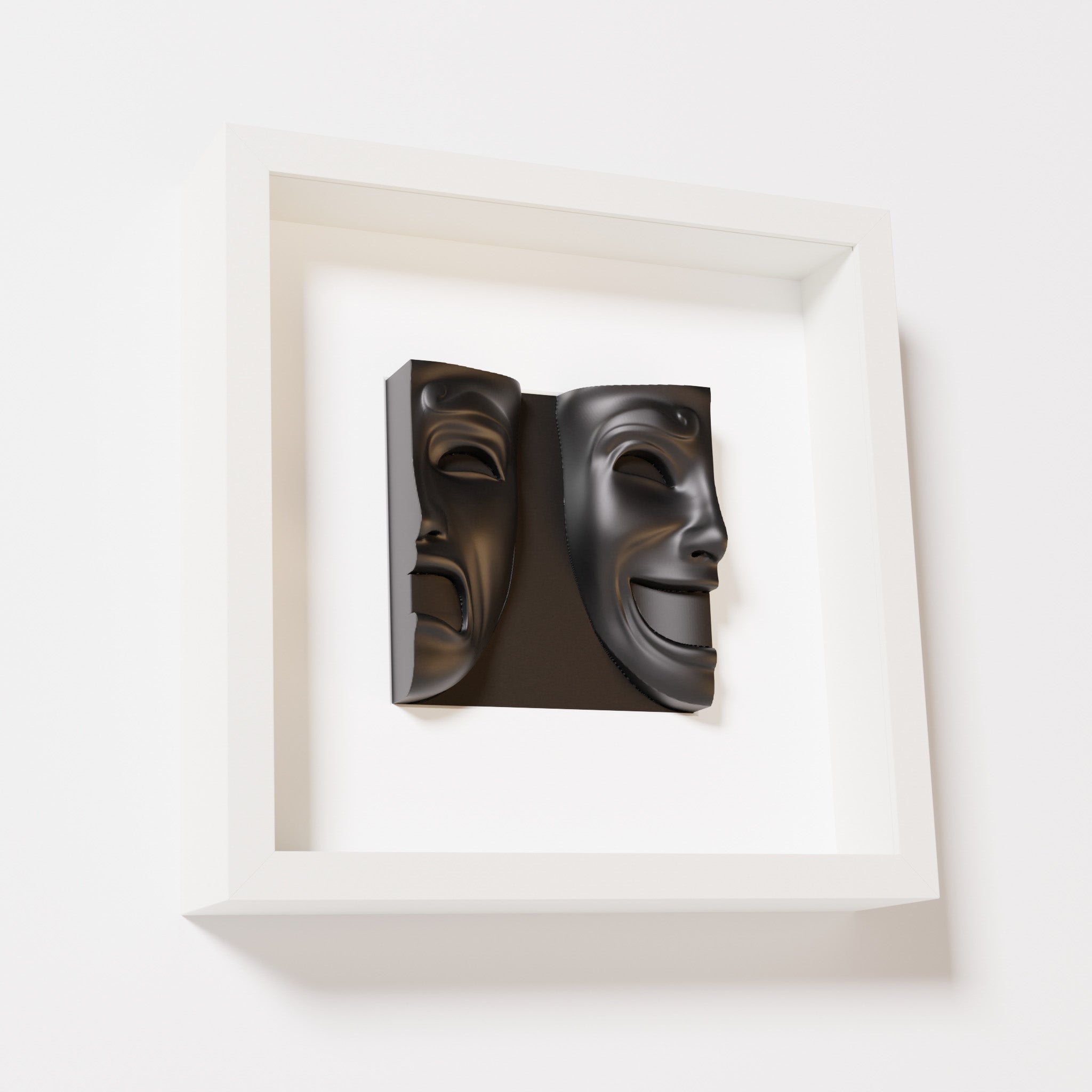 A close-up of a white shadowbox frame containing a matte black sculpture of 'Despair & Joy,' showcasing two expressive masks side by side: one with a sorrowful expression and the other with a joyful smile.