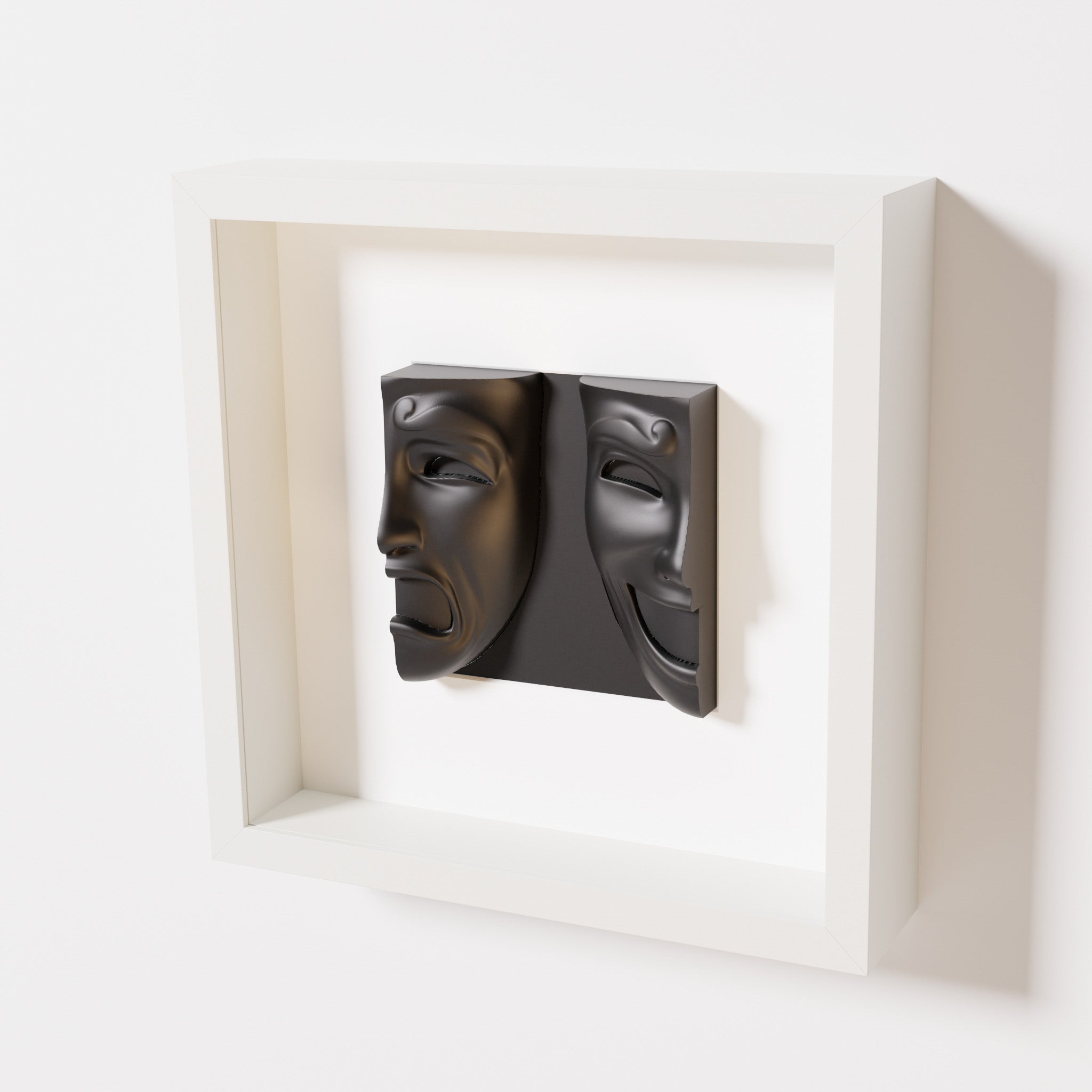 A close-up of a white shadowbox frame containing a matte black sculpture of 'Despair & Joy,' showcasing two expressive masks side by side: one with a sorrowful expression and the other with a joyful smile.