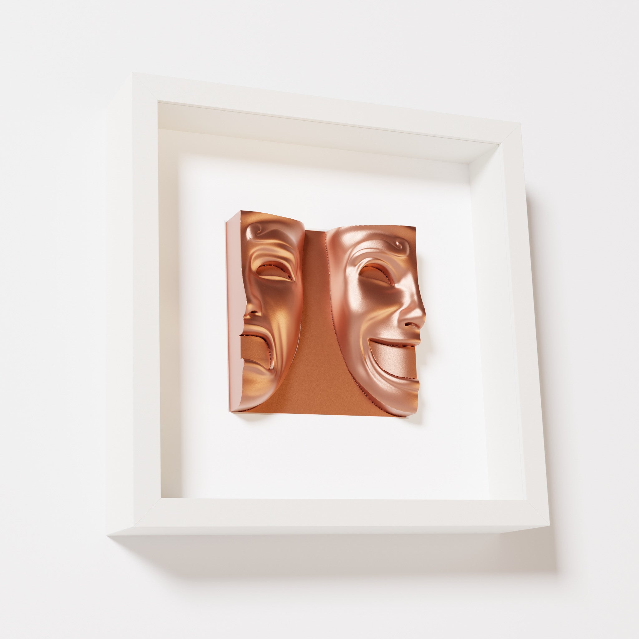 A close-up of a white shadowbox frame containing a chrome copper sculpture of 'Despair & Joy,' showcasing two expressive masks side by side: one with a sorrowful expression and the other with a joyful smile.