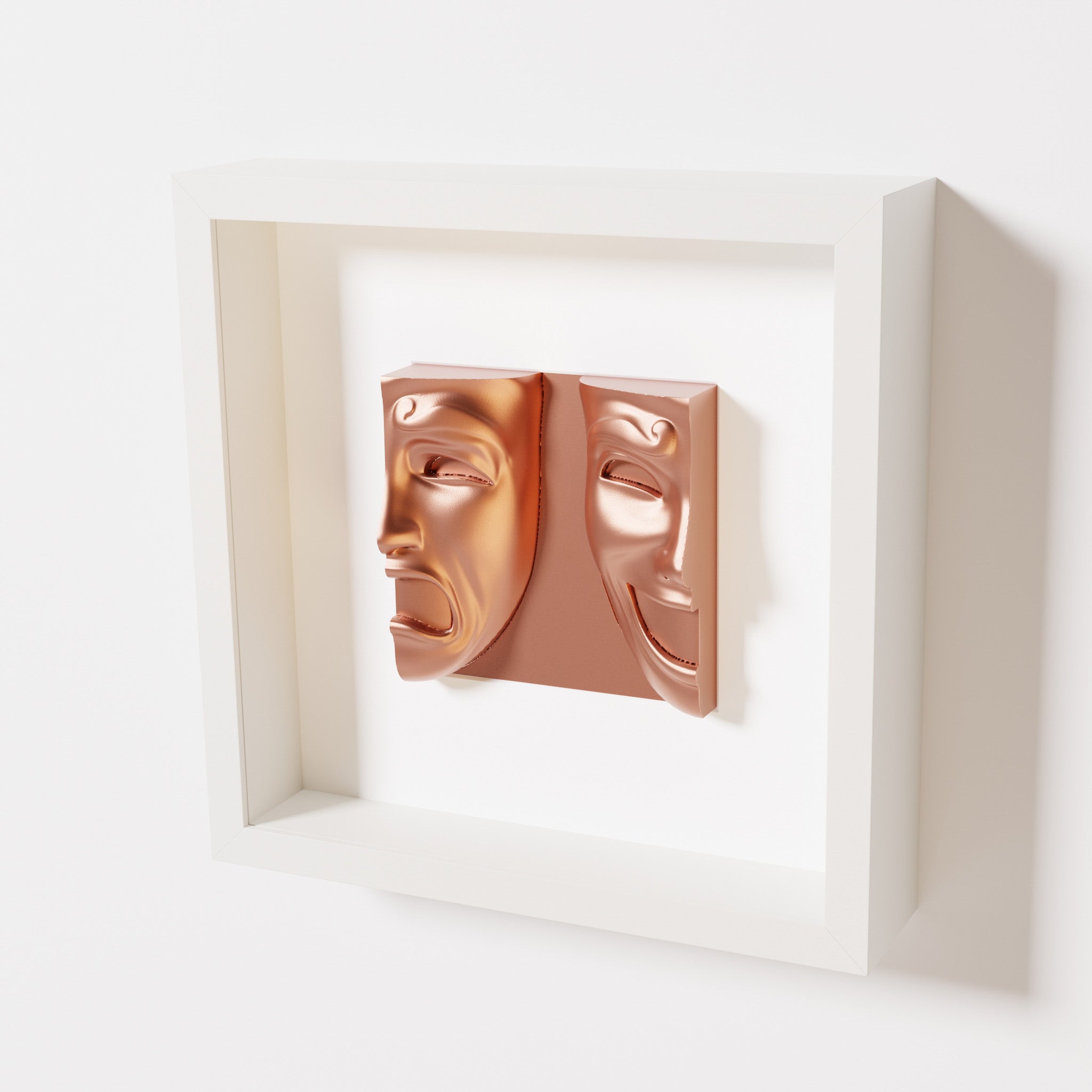 A close-up of a white shadowbox frame containing a chrome copper sculpture of 'Despair & Joy,' showcasing two expressive masks side by side: one with a sorrowful expression and the other with a joyful smile.