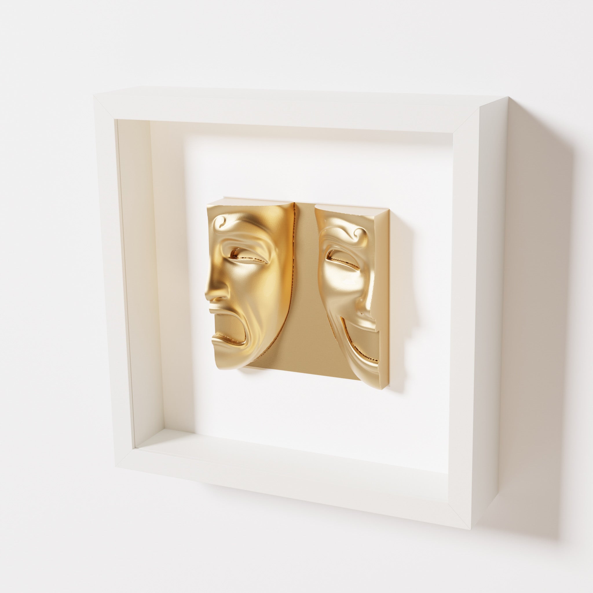 A close-up of a white shadowbox frame containing a chrome gold sculpture of 'Despair & Joy,' showcasing two expressive masks side by side: one with a sorrowful expression and the other with a joyful smile.