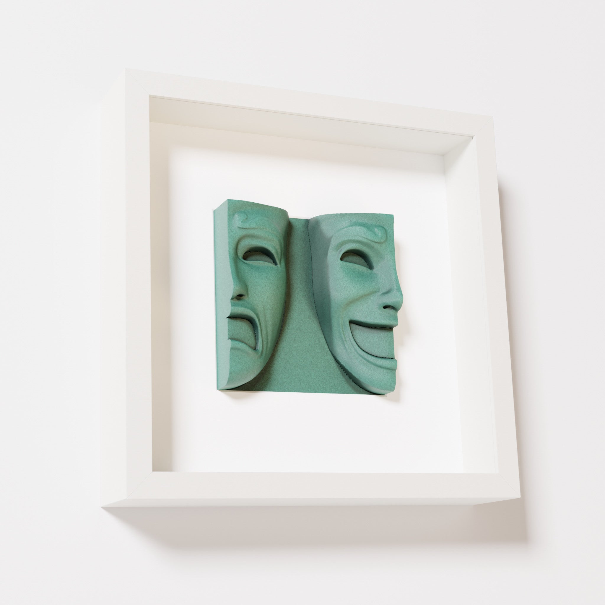 A close-up of a white shadowbox frame containing an old patina sculpture of 'Despair & Joy,' showcasing two expressive masks side by side: one with a sorrowful expression and the other with a joyful smile.