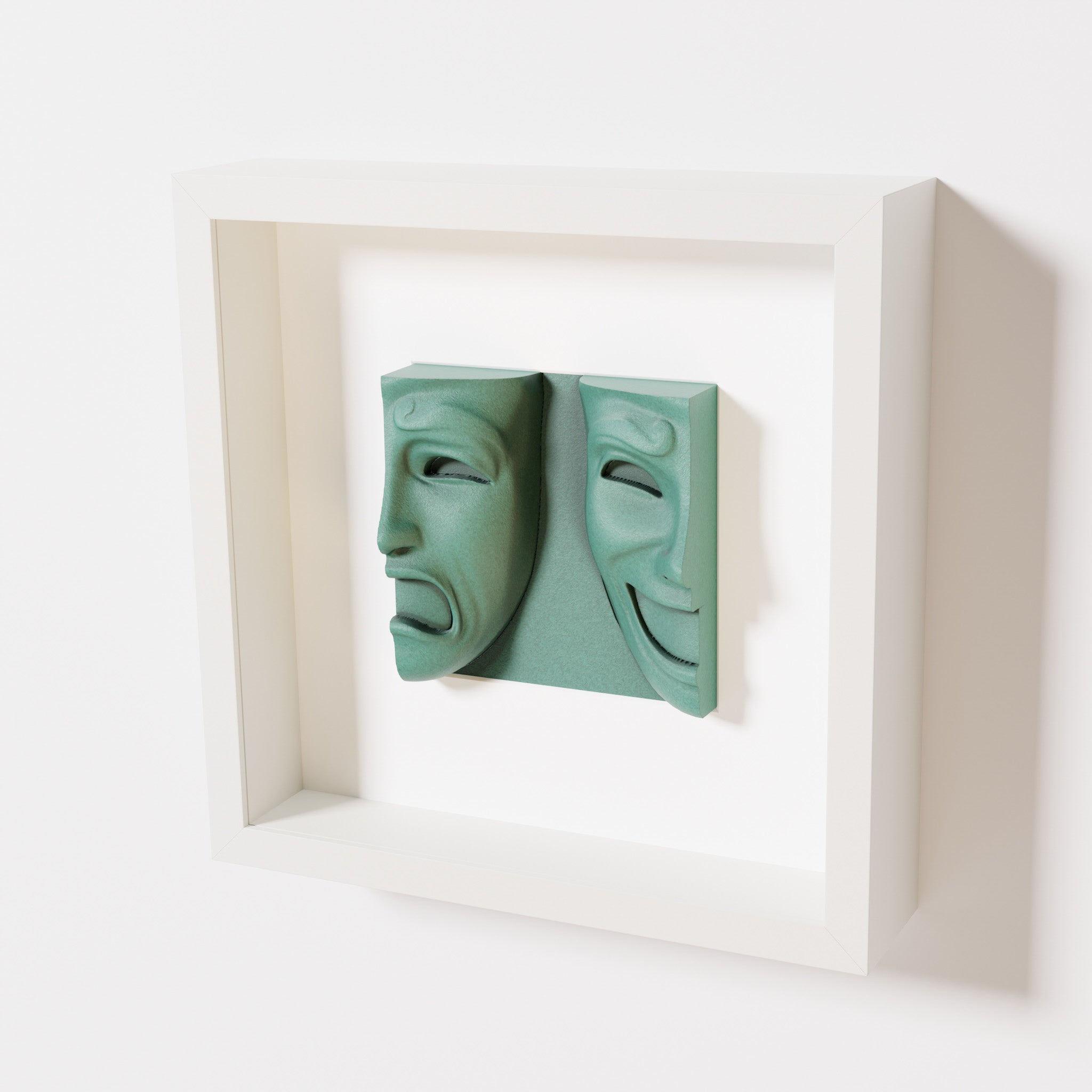 A close-up of a white shadowbox frame containing an old patina sculpture of 'Despair & Joy,' showcasing two expressive masks side by side: one with a sorrowful expression and the other with a joyful smile.