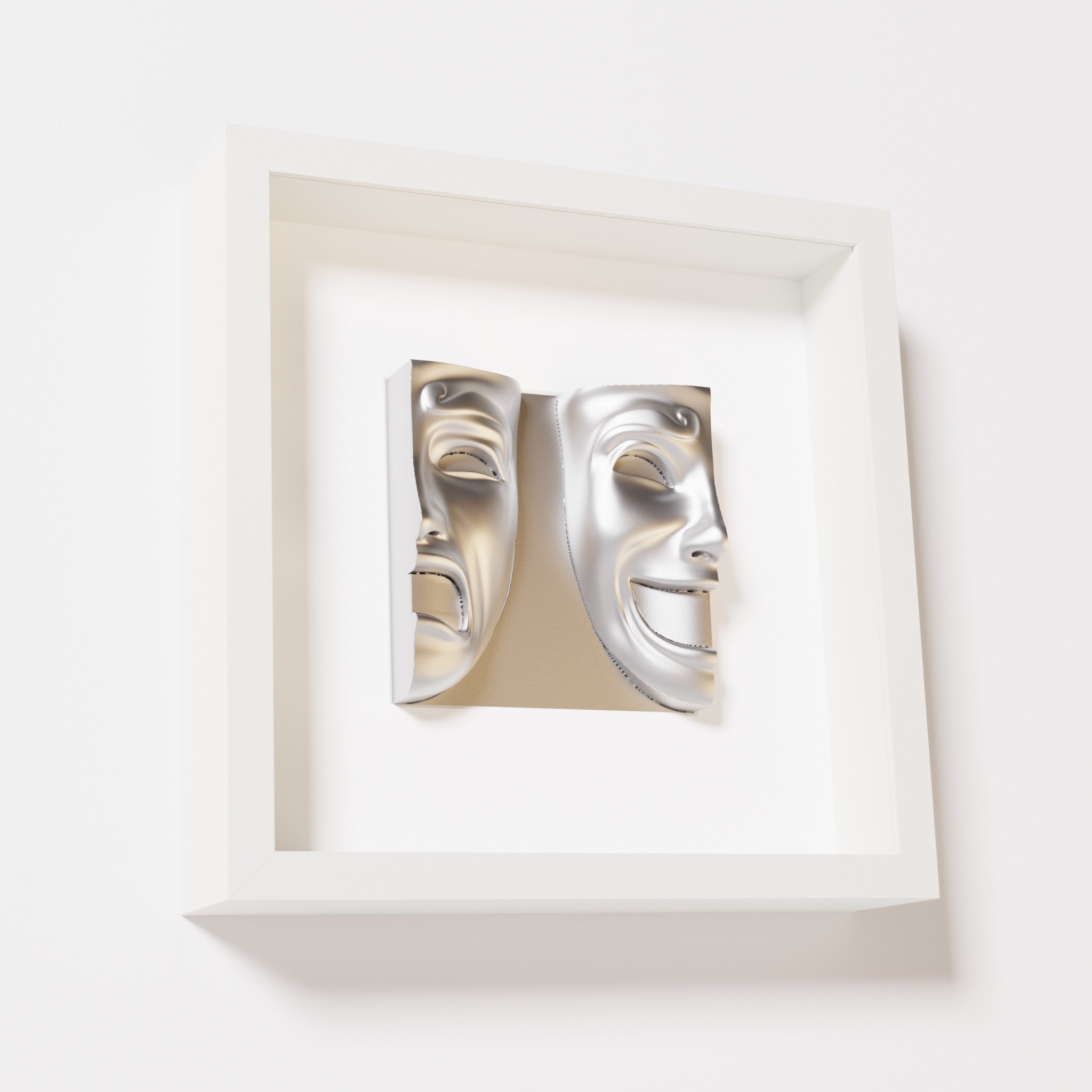 A close-up of a white shadowbox frame containing a chrome silver sculpture of 'Despair & Joy,' showcasing two expressive masks side by side: one with a sorrowful expression and the other with a joyful smile.