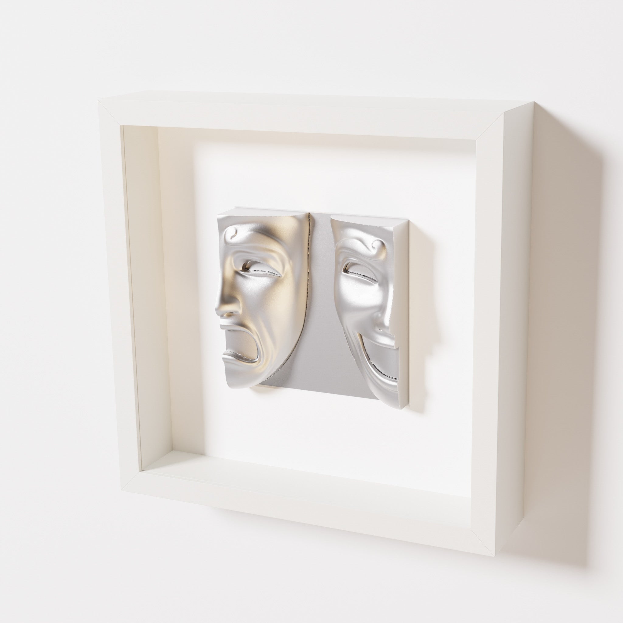 A close-up of a white shadowbox frame containing a chrome silver sculpture of 'Despair & Joy,' showcasing two expressive masks side by side: one with a sorrowful expression and the other with a joyful smile.