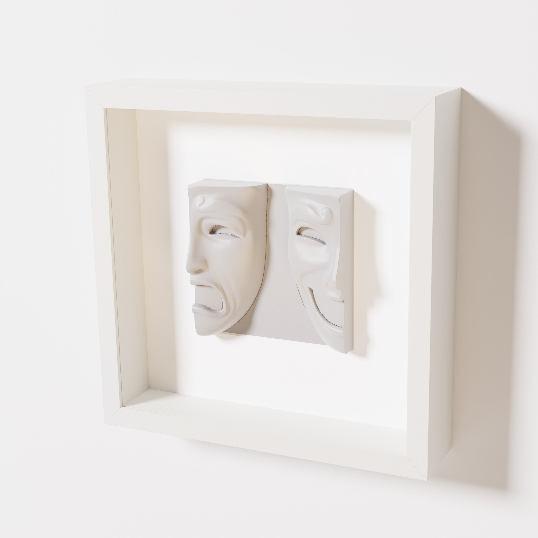 A close-up of a white shadowbox frame containing a marble white sculpture of 'Despair & Joy,' showcasing two expressive masks side by side: one with a sorrowful expression and the other with a joyful smile.