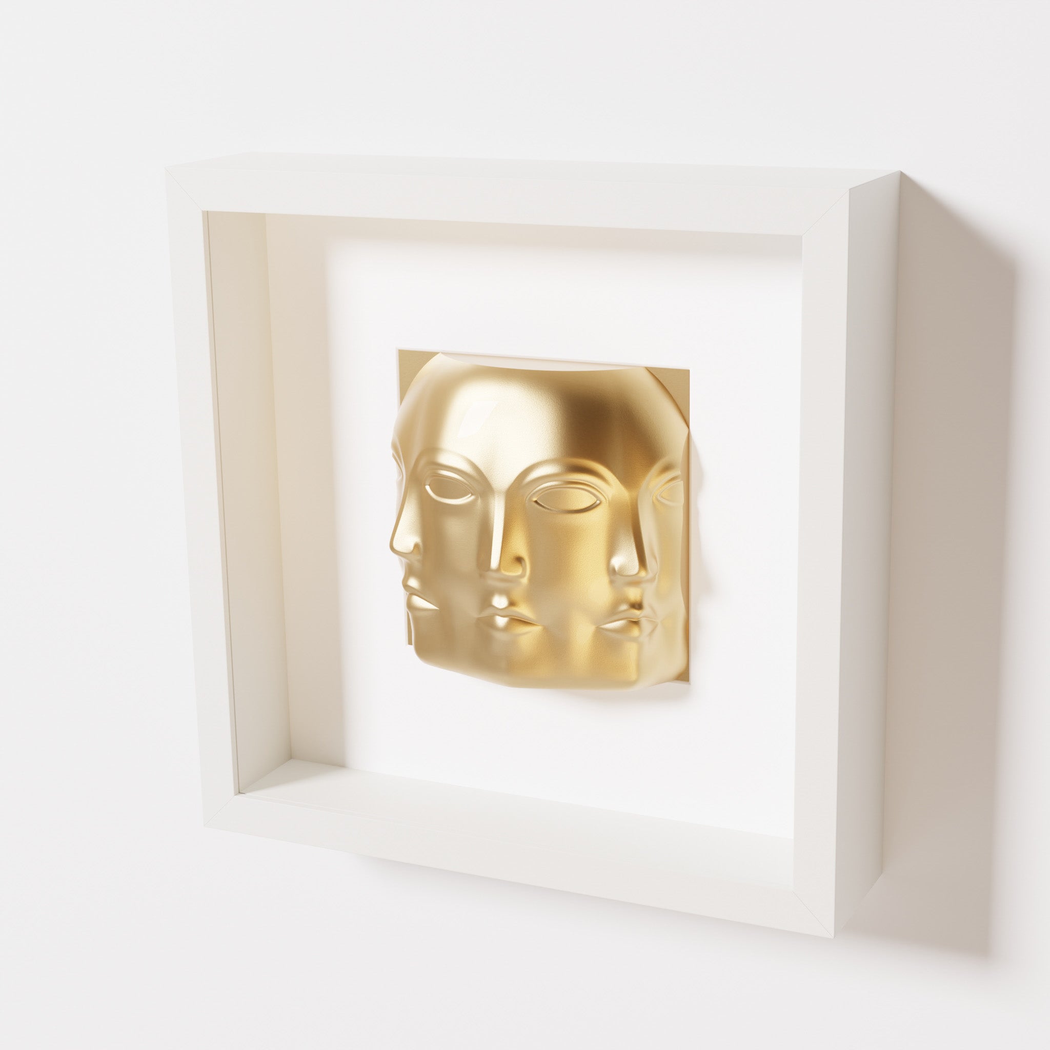 A close-up of a white shadowbox frame containing a chrome gold sculpture of 'Dora,' featuring three interconnected faces merging at the edges, creating a surreal and multi-dimensional effect.