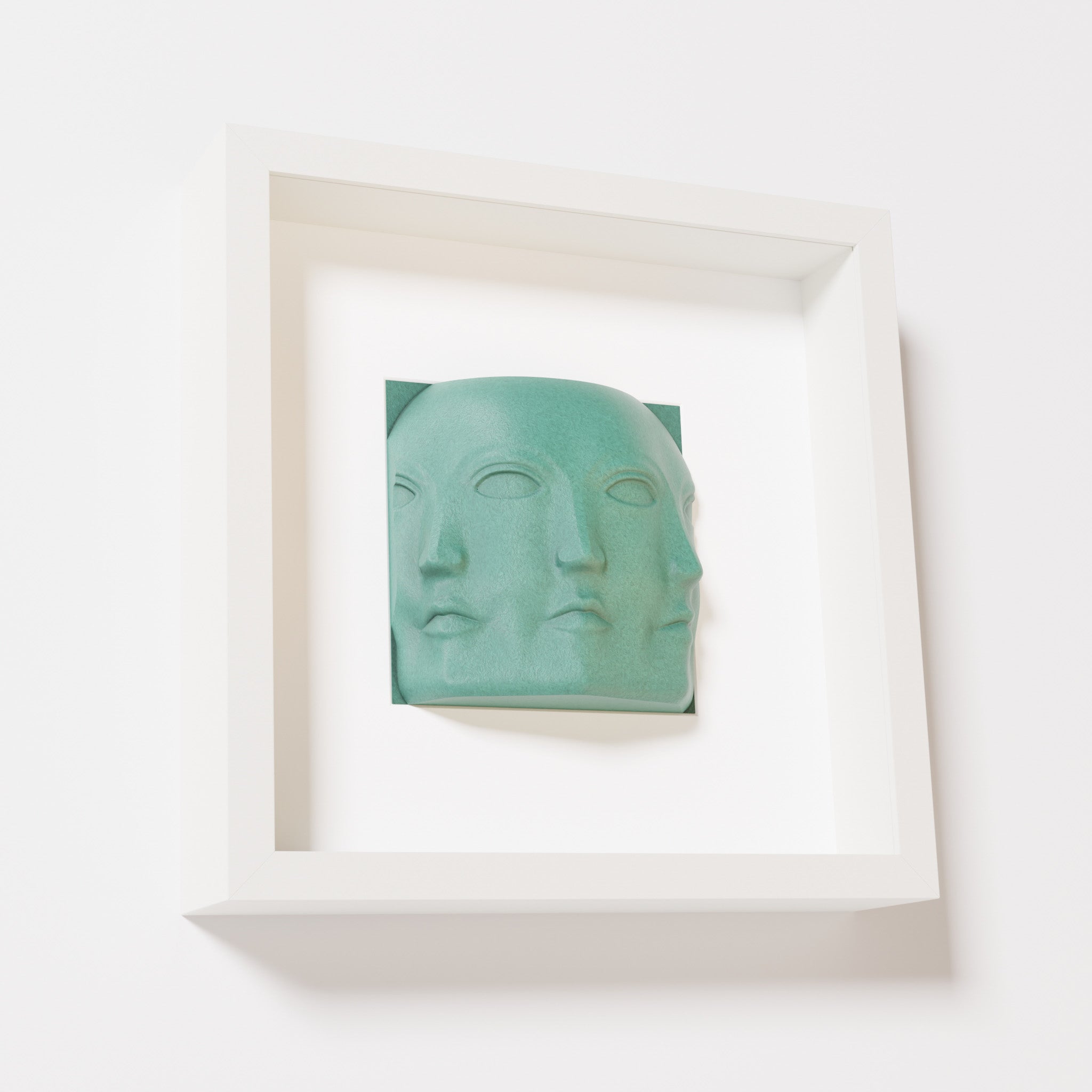 A close-up of a white shadowbox frame containing an old patina sculpture of 'Dora,' featuring three interconnected faces merging at the edges, creating a surreal and multi-dimensional effect.