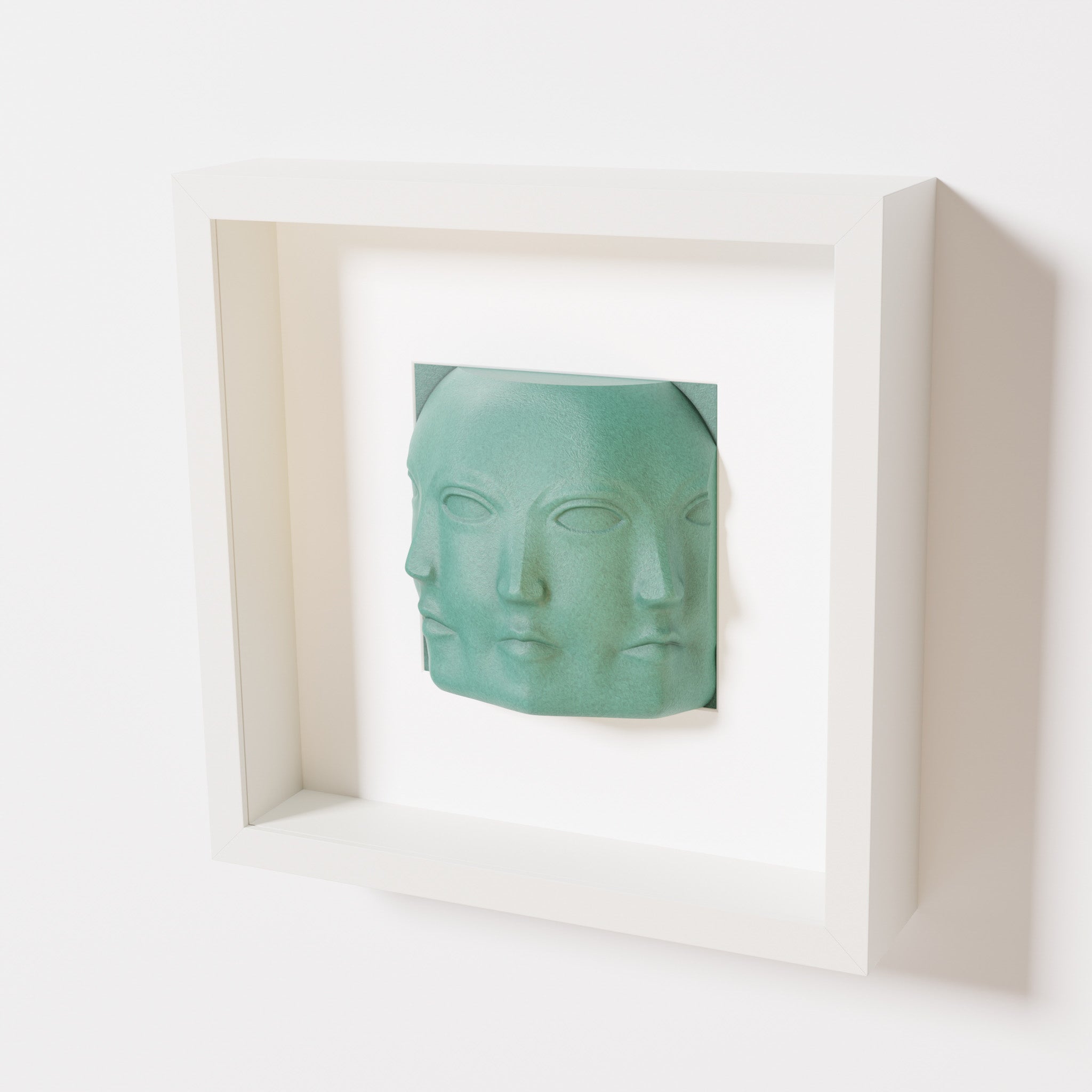 A close-up of a white shadowbox frame containing an old patina sculpture of 'Dora,' featuring three interconnected faces merging at the edges, creating a surreal and multi-dimensional effect.