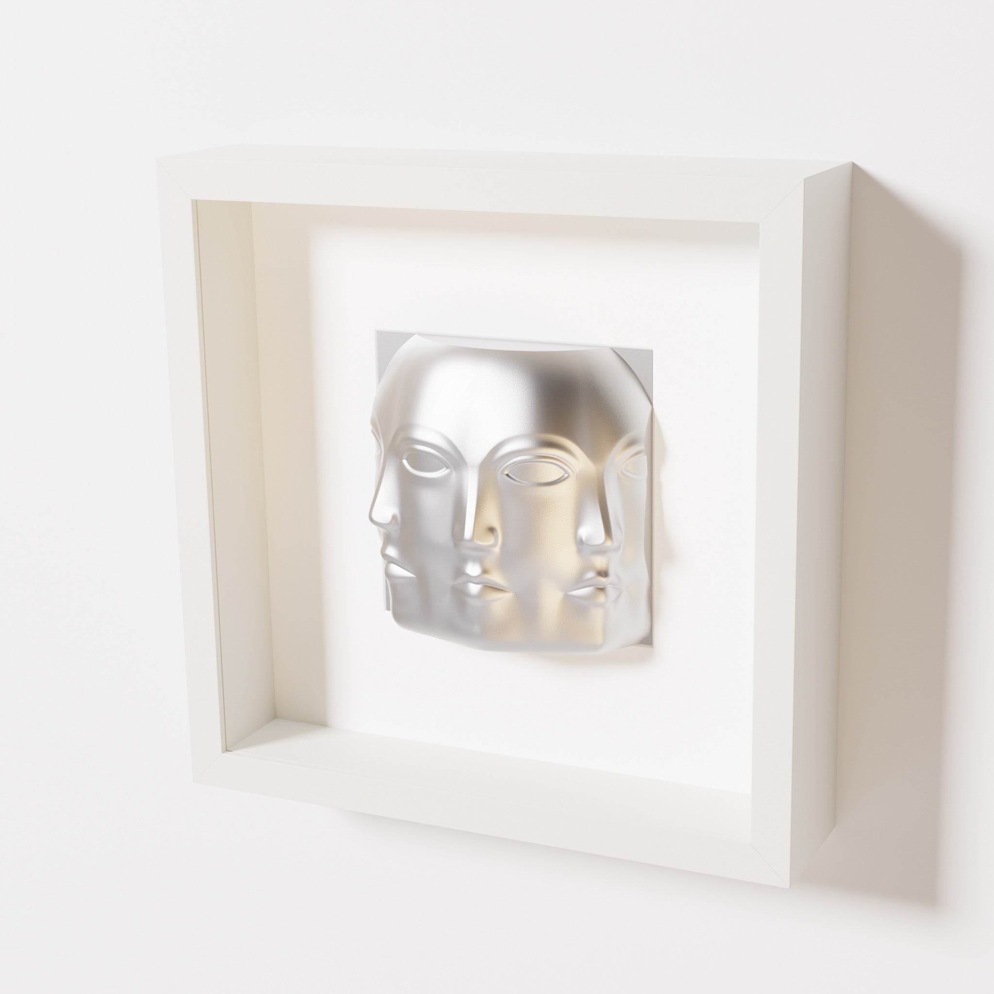 A close-up of a white shadowbox frame containing a chrome silver sculpture of 'Dora,' featuring three interconnected faces merging at the edges, creating a surreal and multi-dimensional effect.