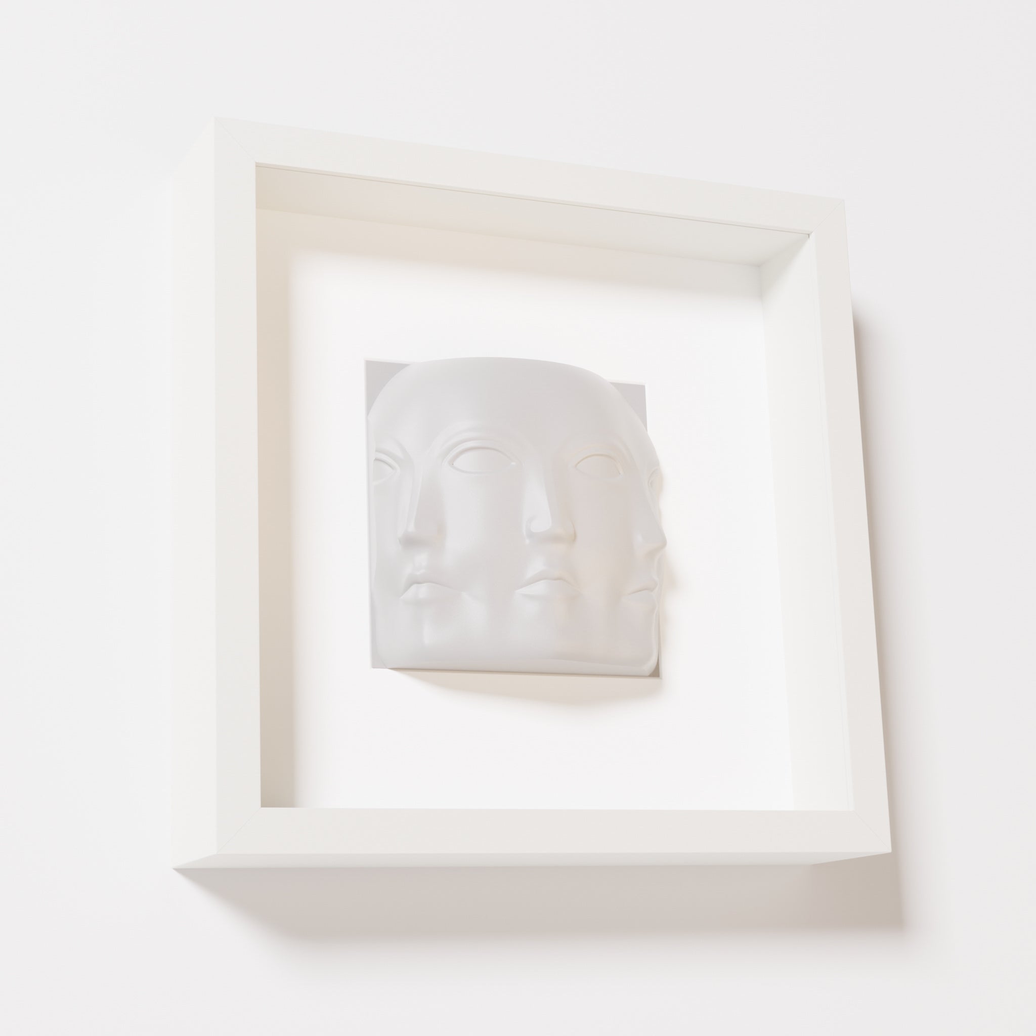 A close-up of a white shadowbox frame containing a marble white sculpture of 'Dora,' featuring three interconnected faces merging at the edges, creating a surreal and multi-dimensional effect.