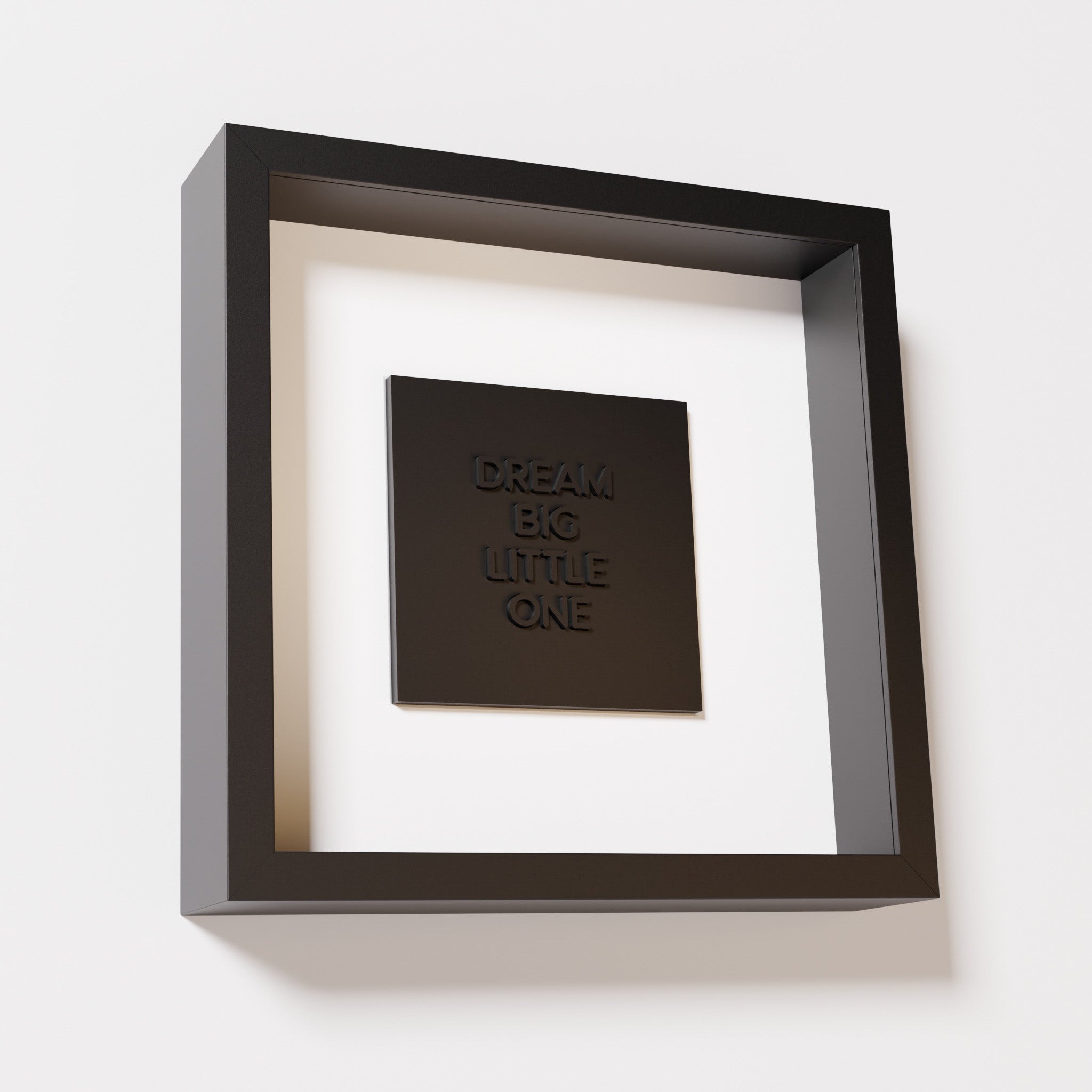 A close-up of a black shadowbox frame containing a matte black artwork with the phrase 'Dream big little one,' showcasing bold, raised lettering on a smooth background.