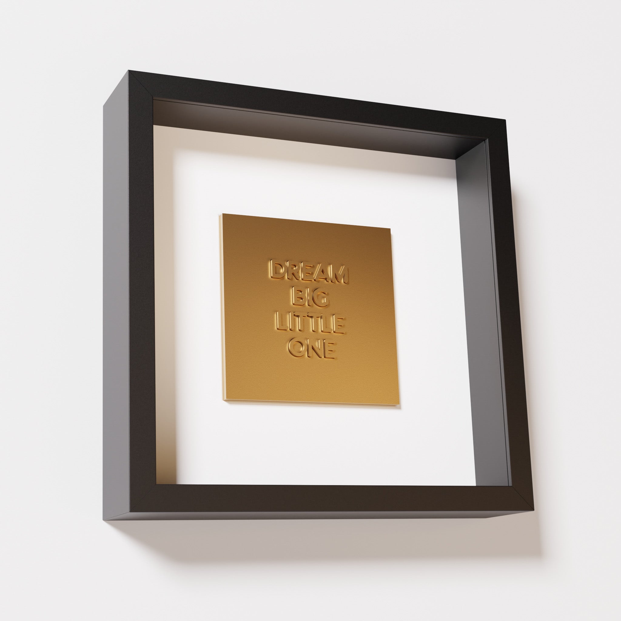 A close-up of a black shadowbox frame containing a chrome gold artwork with the phrase 'Dream big little one,' showcasing bold, raised lettering on a reflective golden surface.