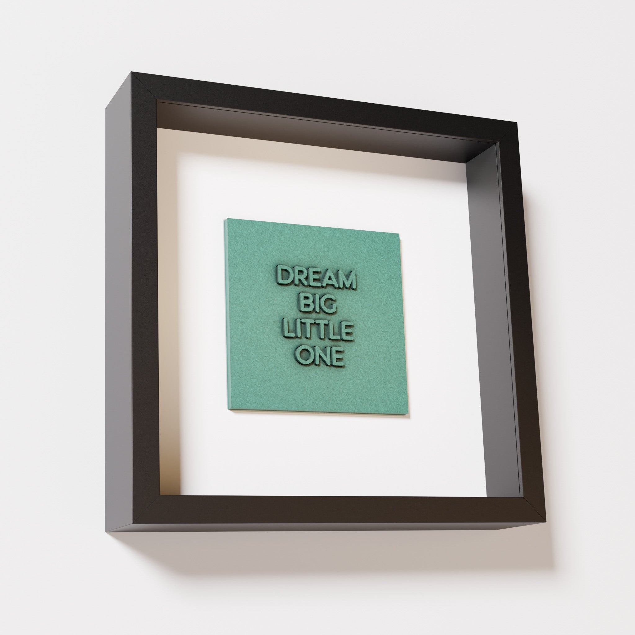 A close-up of a black shadowbox frame containing an old patina artwork with the phrase 'Dream big little one,' showcasing bold, raised lettering on a textured green surface.